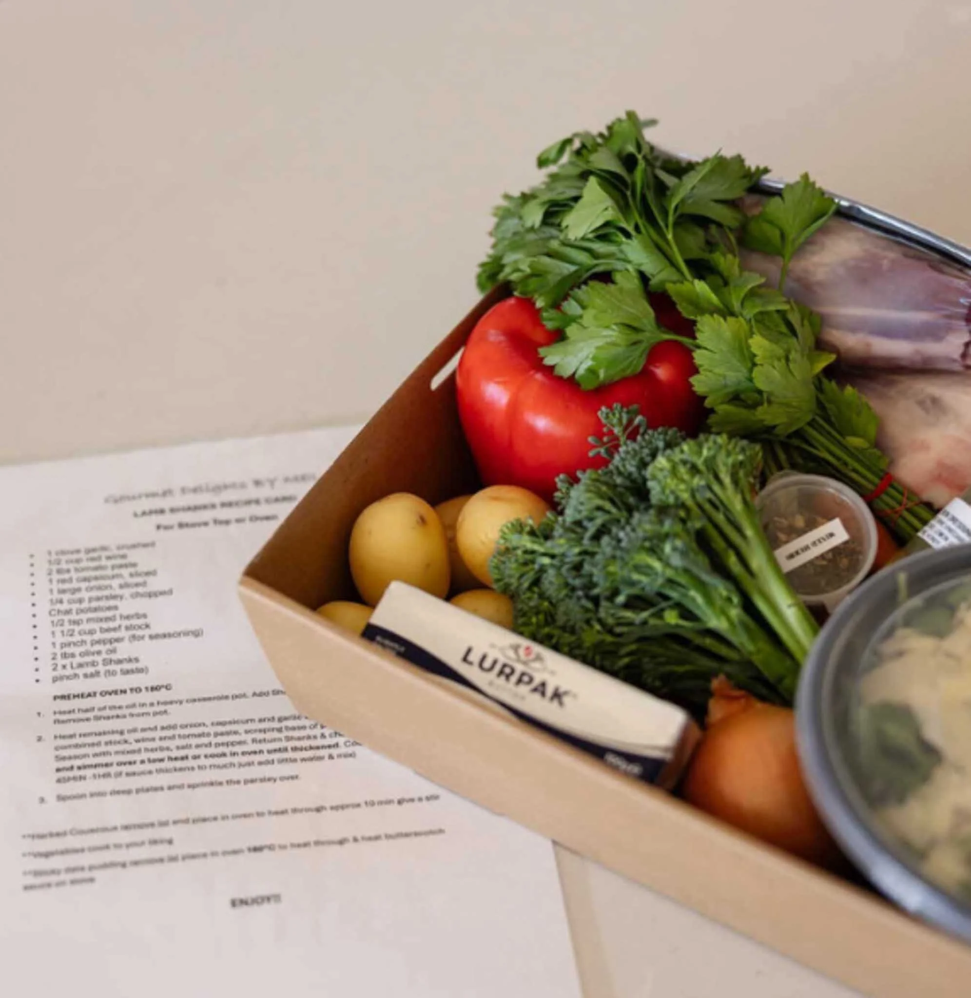 A box of fresh vegetables including a red tomato, potatoes, leafy greens, and herbs, with a partially visible printed recipe below.