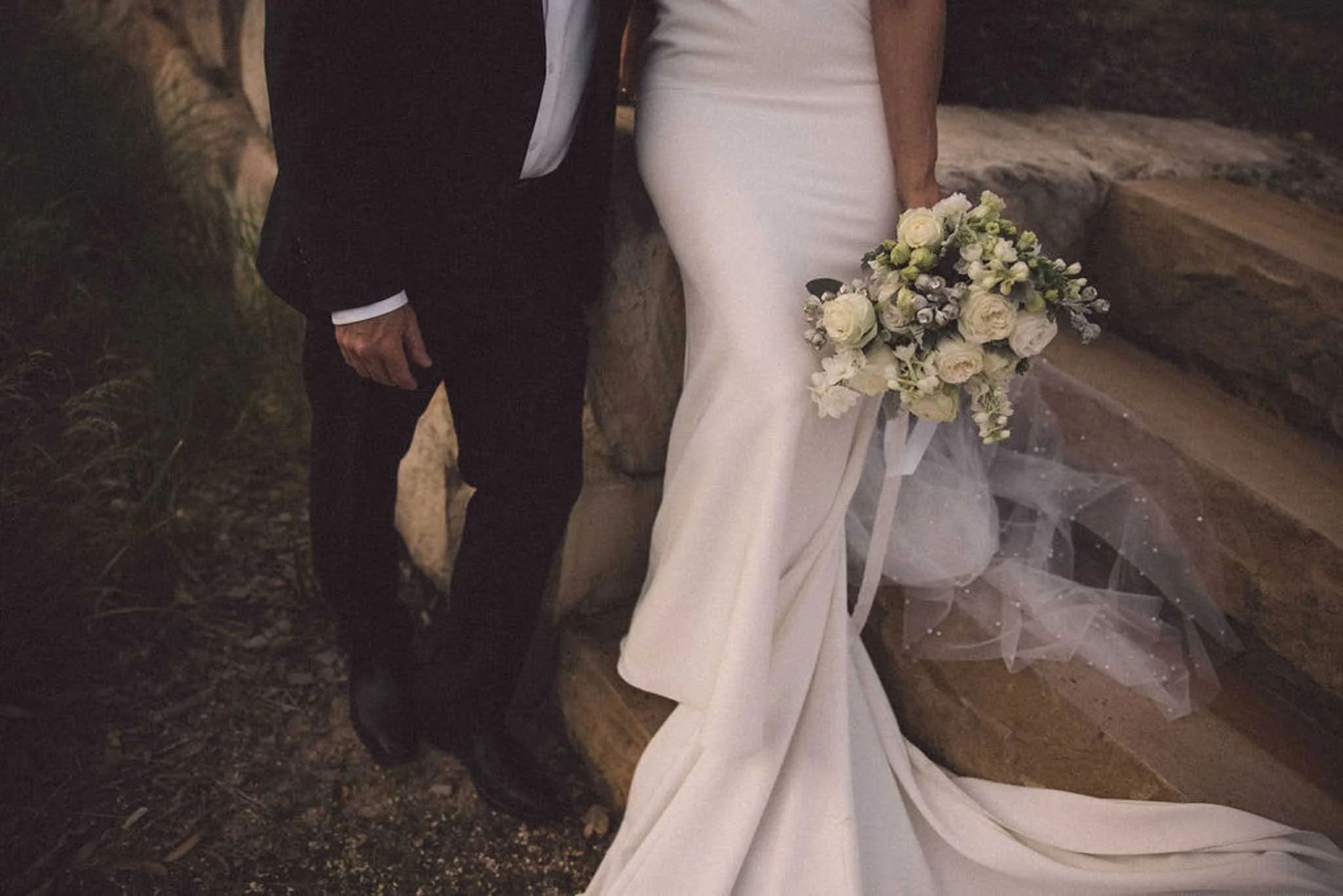 Couple standing outdoors at night, the woman in a white wedding dress holding a bouquet of white flowers, the man in a dark suit with a white shirt and a black vest.