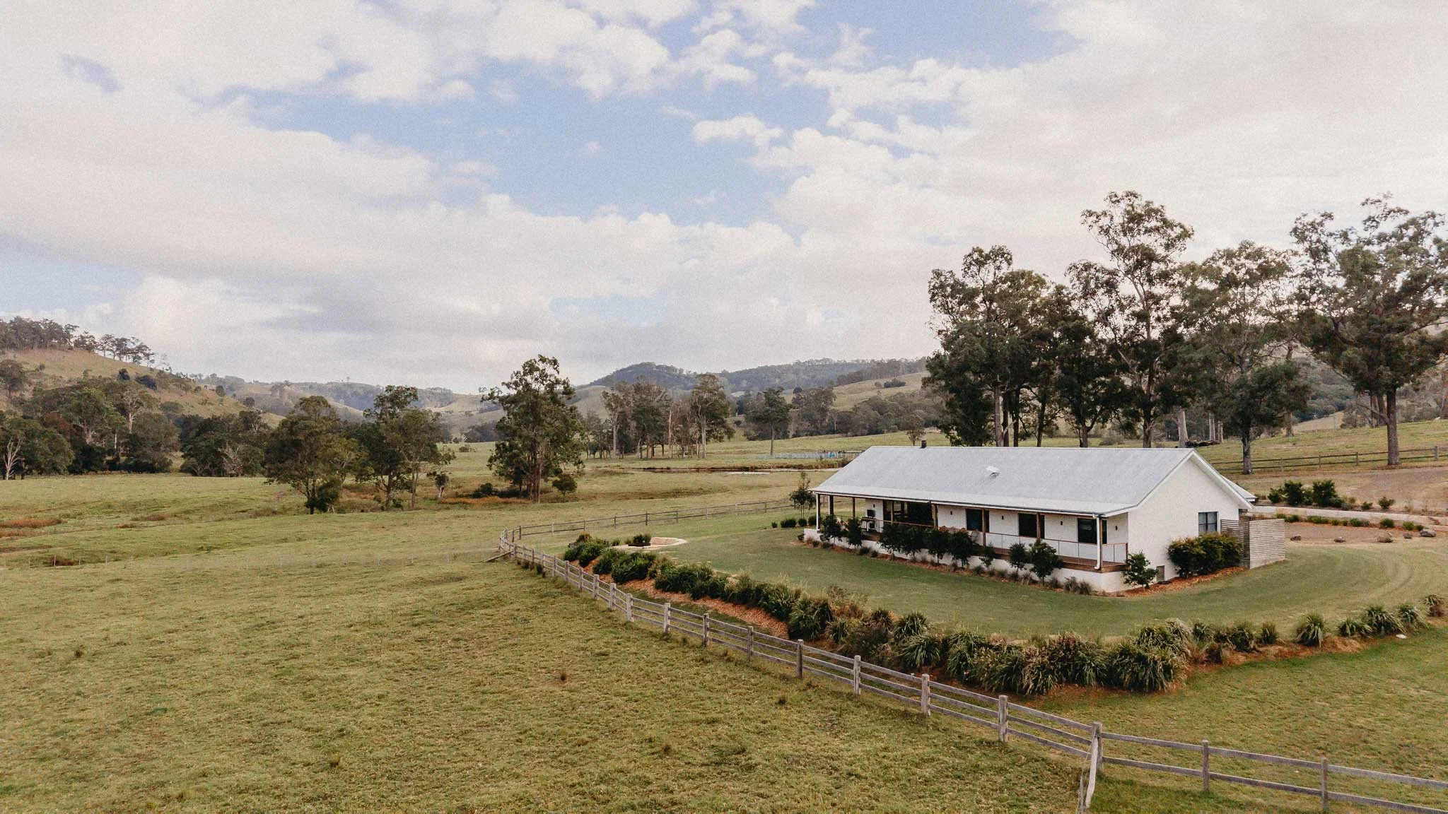 A rural landscape with rolling hills, trees, a white house with a gray roof, and a fenced yard under a partly cloudy sky.