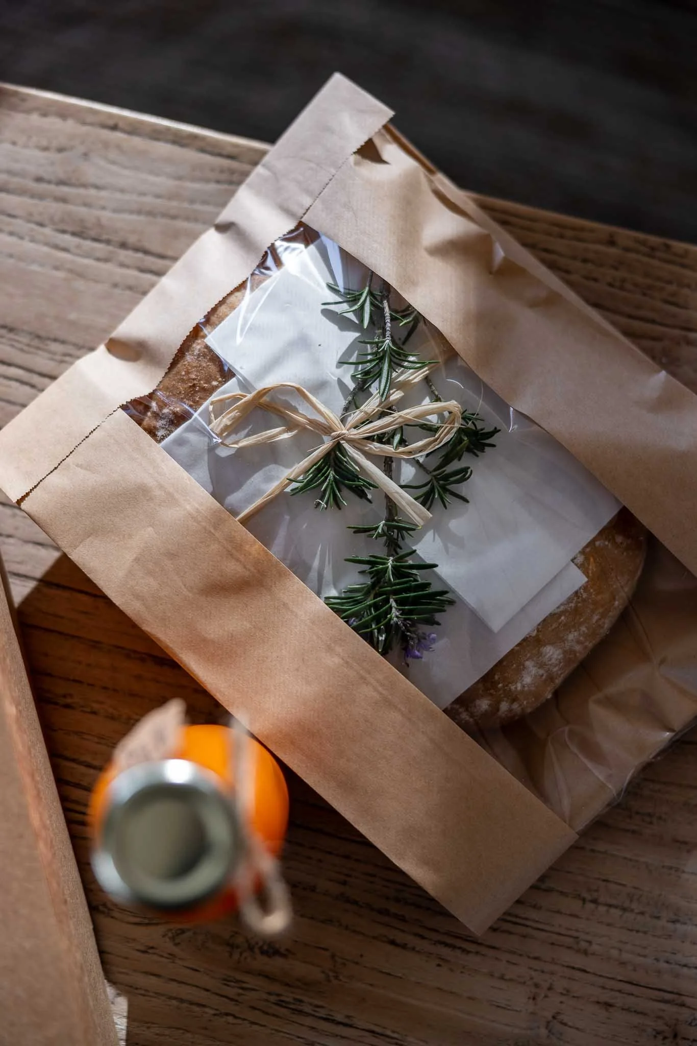 A brown paper bag with a transparent window revealing a sprig of rosemary inside, placed on a wooden surface, with a blurry orange object and a silver can top in the foreground.