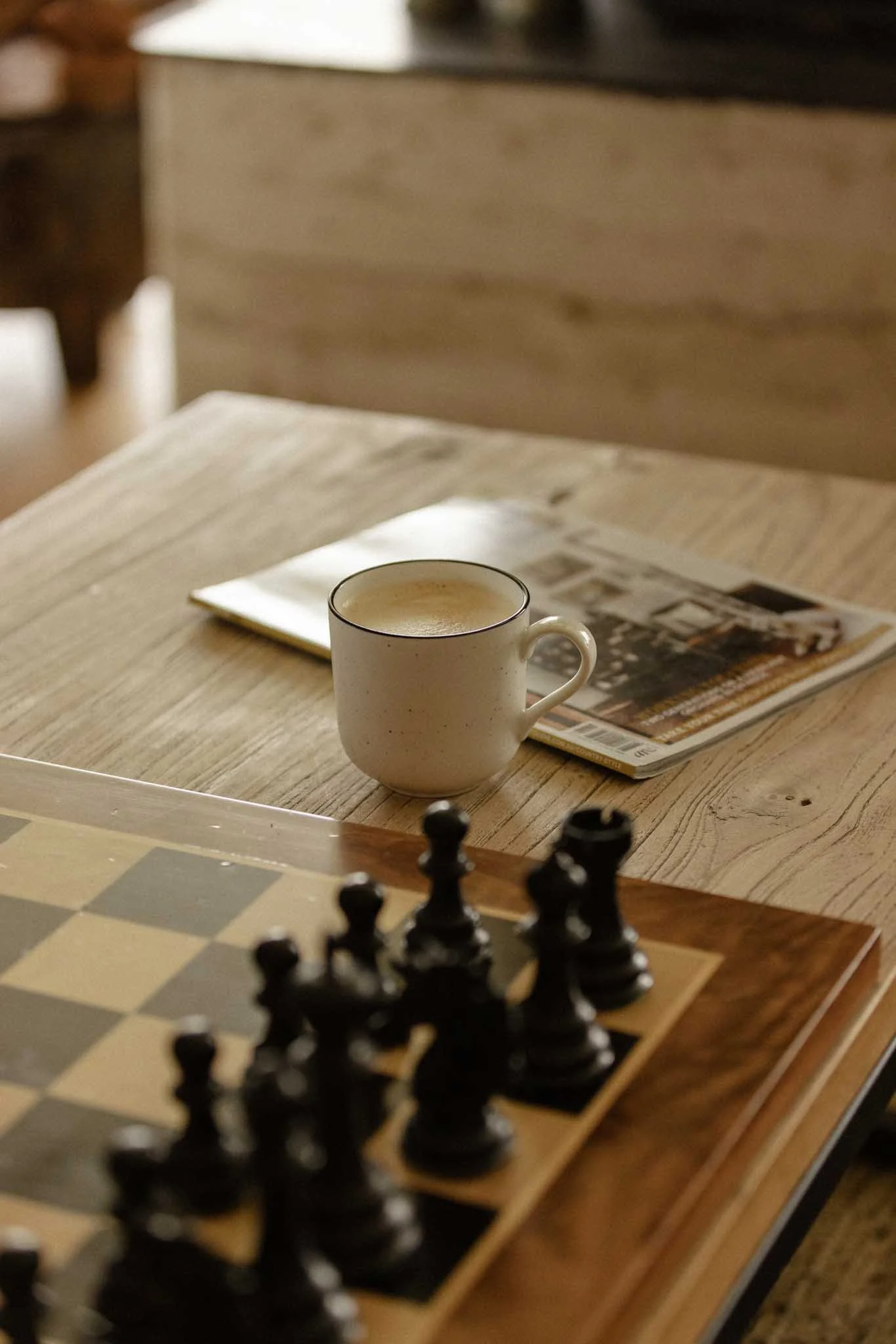 A wooden table with a chessboard, a white coffee mug filled with a hot beverage, and a magazine or newspaper. The background features a blurred room with wooden furniture.