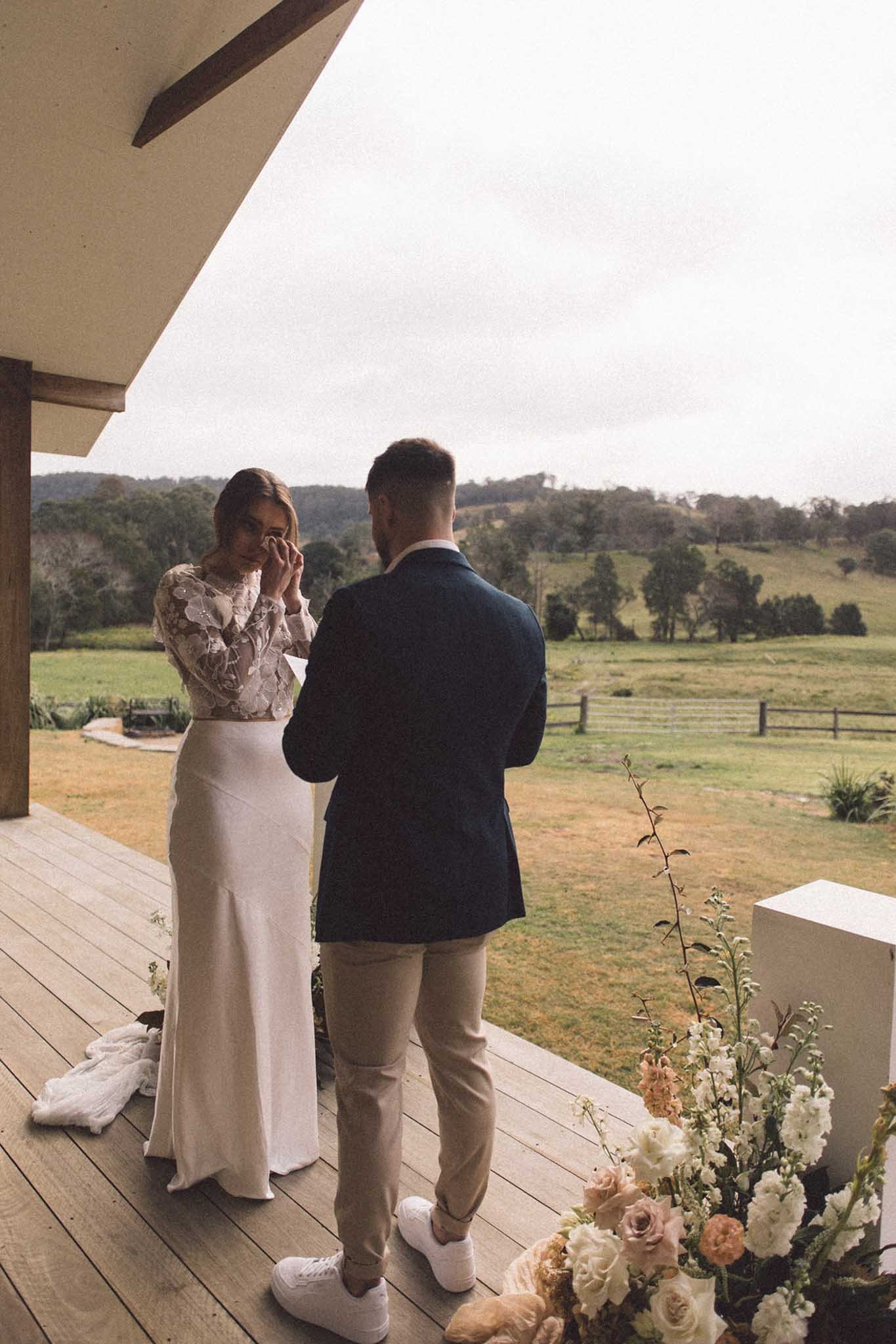 A couple having a wedding ceremony outdoors on a deck overlooking a greenfield with trees and hills in the background. The woman is adjusting her veil, wearing a white lace dress, and the man is dressed in a navy blazer, light-colored pants, and sneakers.