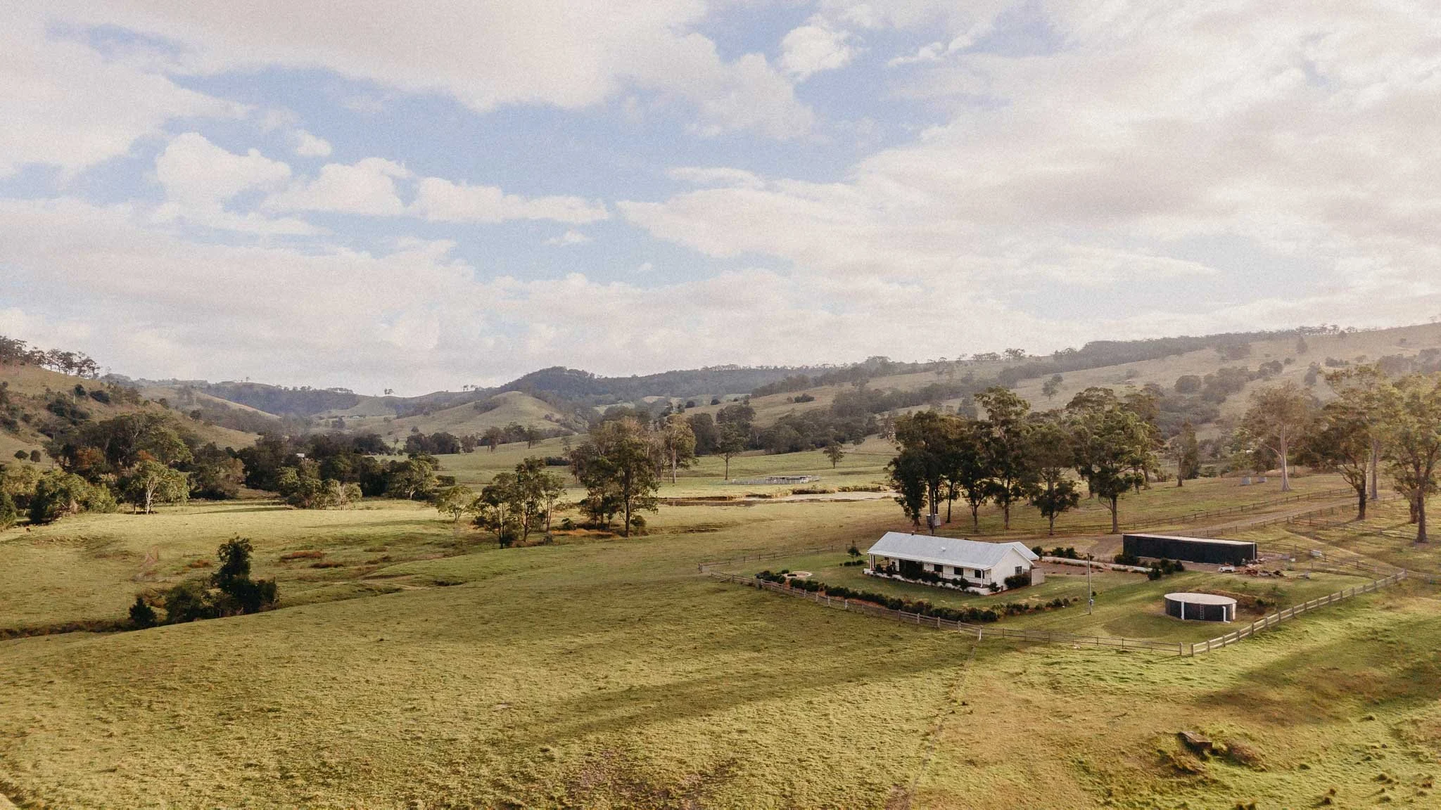 A rural landscape with rolling green hills, scattered trees, a white house with a porch, and a fenced yard. In the background, there are more hills and a partly cloudy sky.