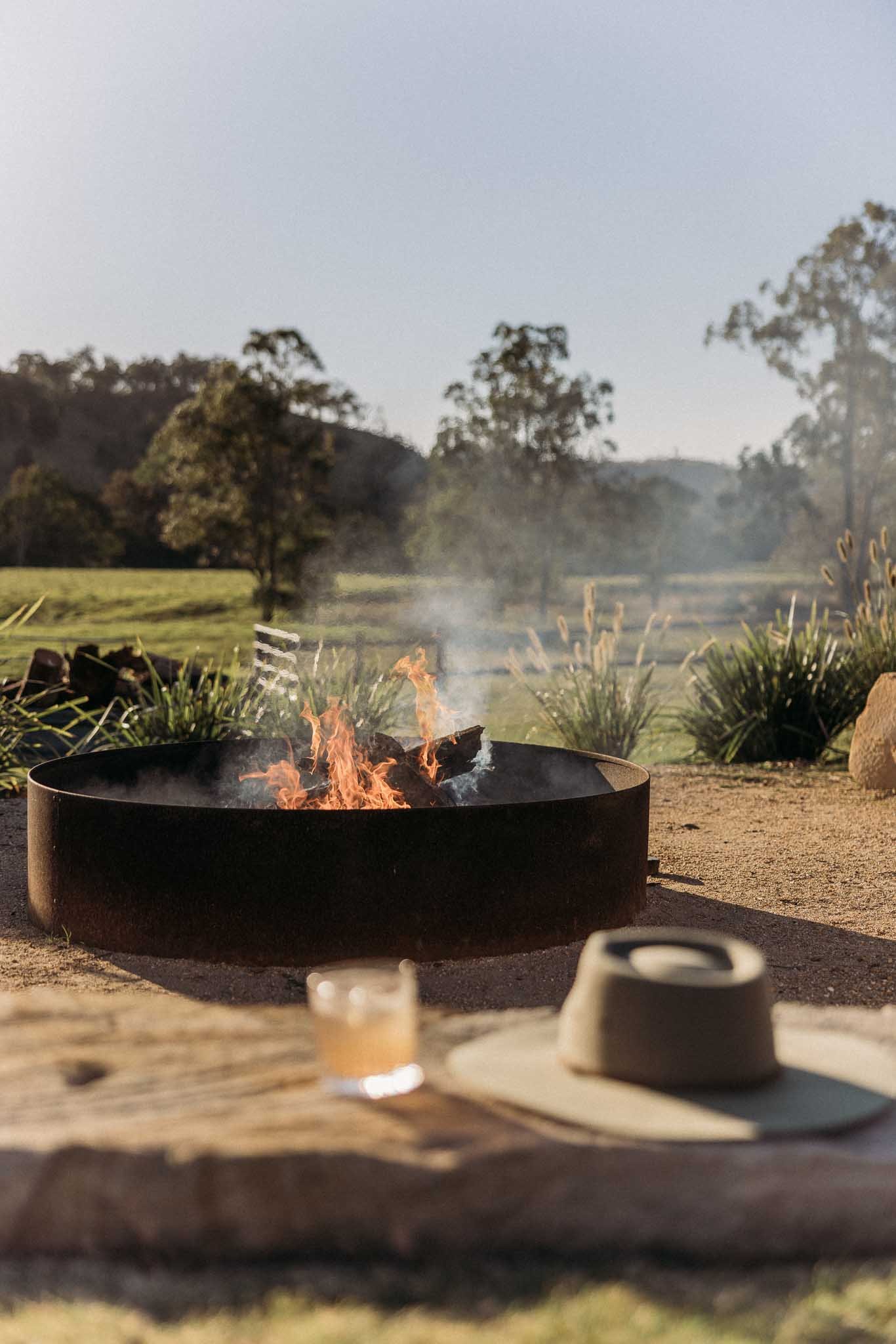 Fire pit burning outdoors with a background of trees and a clear sky, with blurred cups and a table in the foreground.