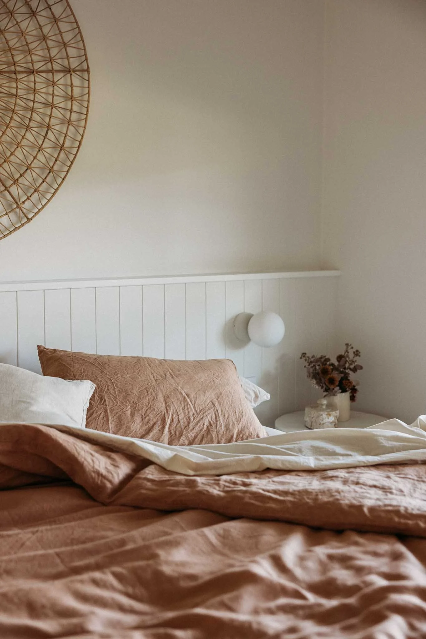 A cozy bedroom with a bed covered in peach-colored bedding, pillows, and a beige blanket. There is a white nightstand with a marble lamp and a vase of dried flowers. The wall behind the bed has white paneling and a round wall light. A woven decorative piece is partially visible on the upper left.