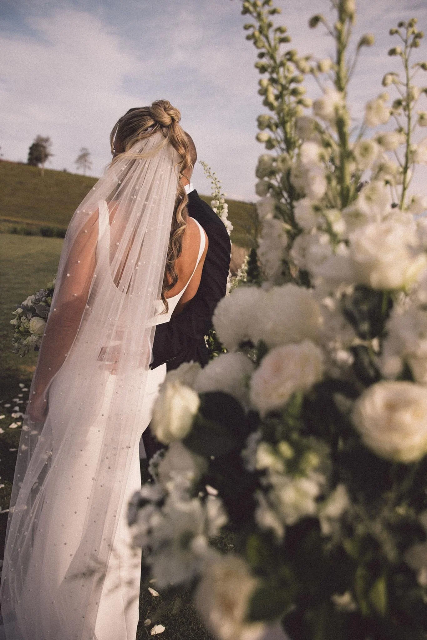 A bride and groom embracing outdoors, with the bride wearing a veil and white dress, and the groom in a dark suit, surrounded by white flowers and a grassy hillside in the background.