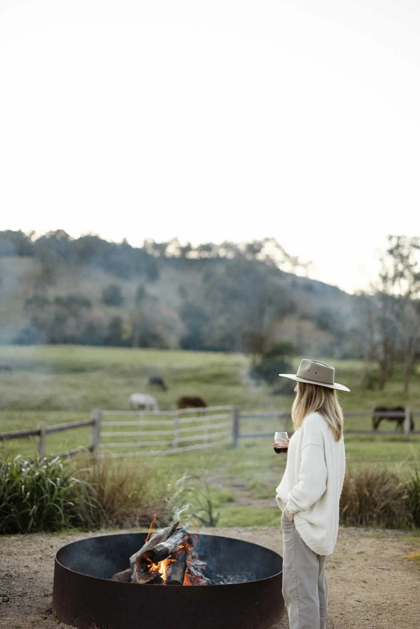 A woman with blonde hair wearing a beige hat and white oversized sweater stands near a fire pit, holding a glass of red wine, with a pastoral landscape and grazing cows in the background.