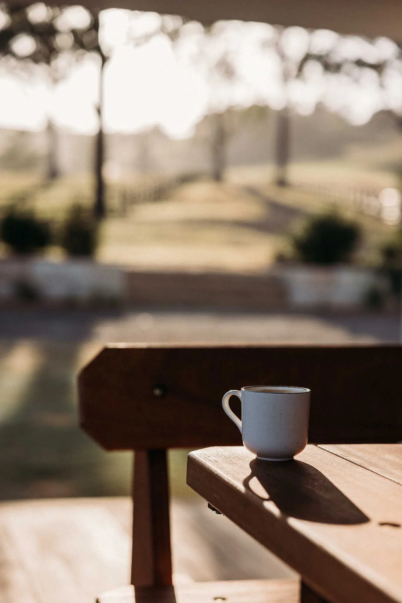 A white coffee mug sitting on a wooden picnic table outside, with a blurred background of sunlight filtering through trees.