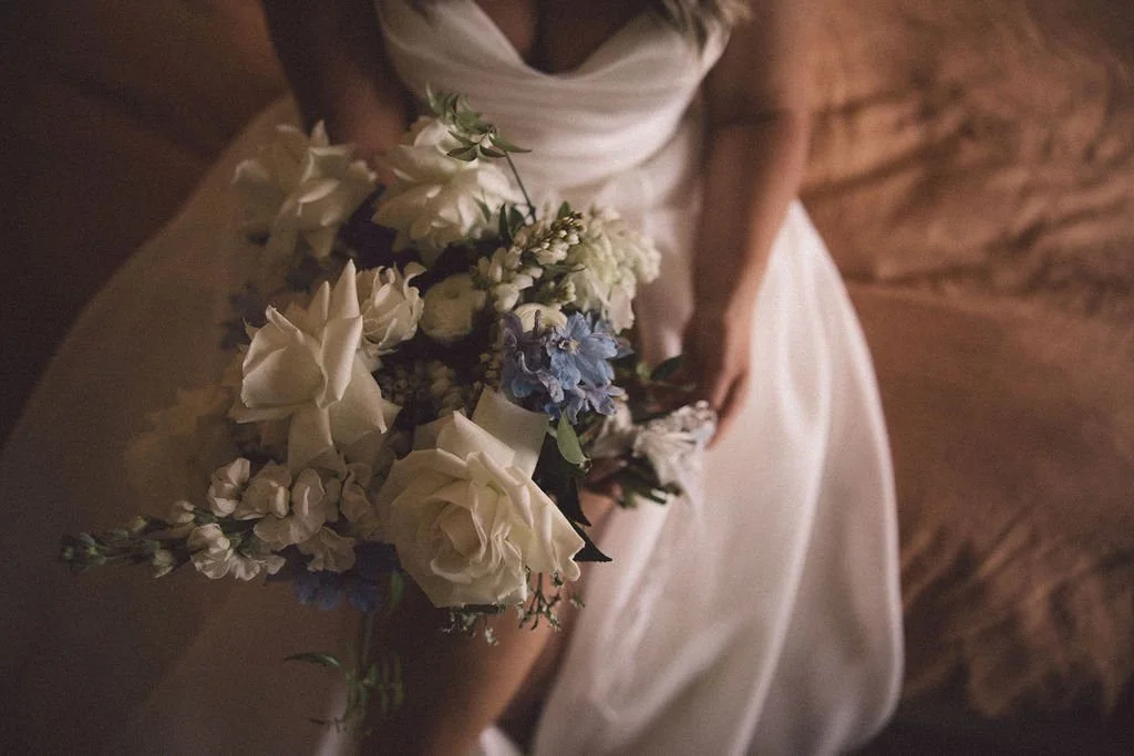 A woman in a wedding dress holding a bouquet of white and blue flowers in a rustic, wood-paneled setting.