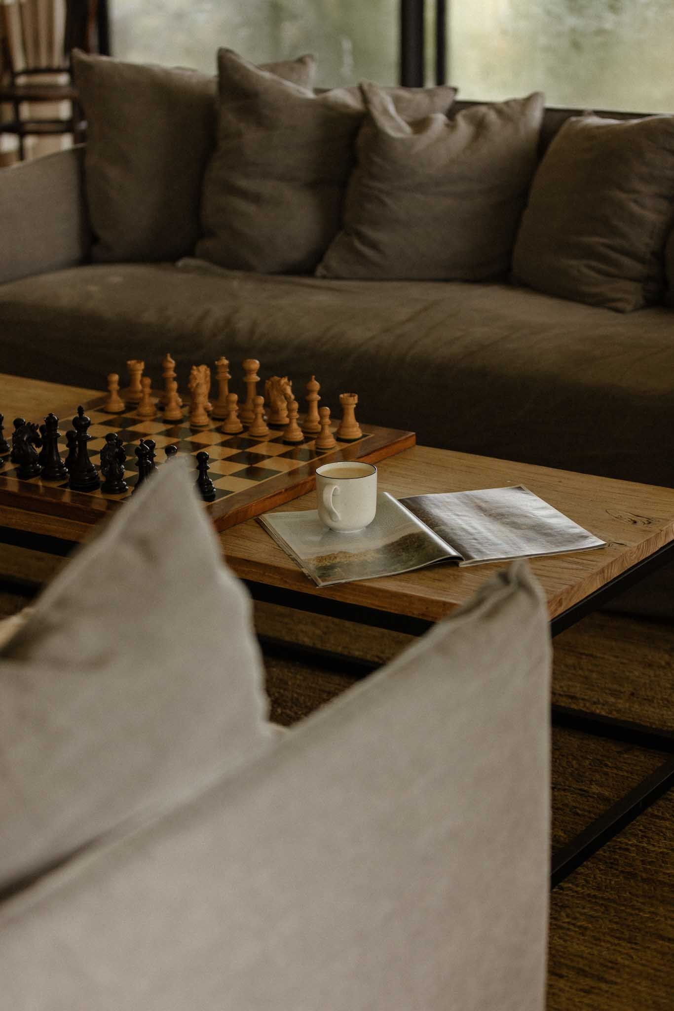 A cozy living room scene with a brown sofa and cushions, a wooden coffee table with a chessboard, a cup of coffee, and an open magazine, near large windows with blurred outdoor view