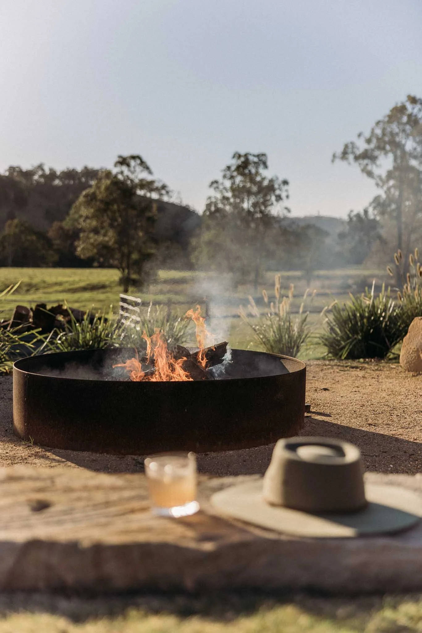 A daytime outdoor scene with a fire pit in the foreground, some small objects on a table, and a scenic background of trees, hills, and clear sky.
