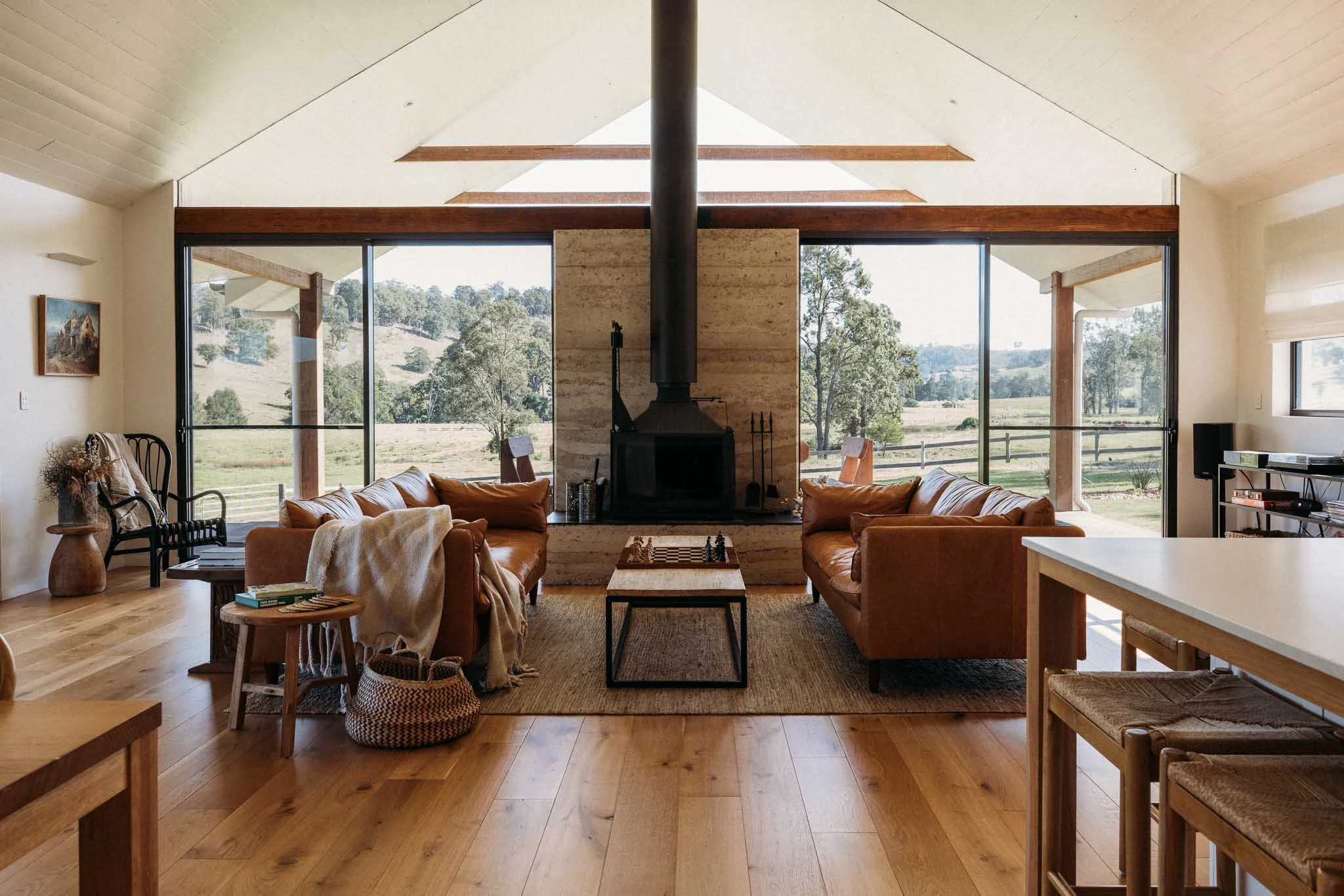 Living room with large windows showing outdoor landscape, tan sofas, a central fireplace, wooden floor, and various chairs and decor.