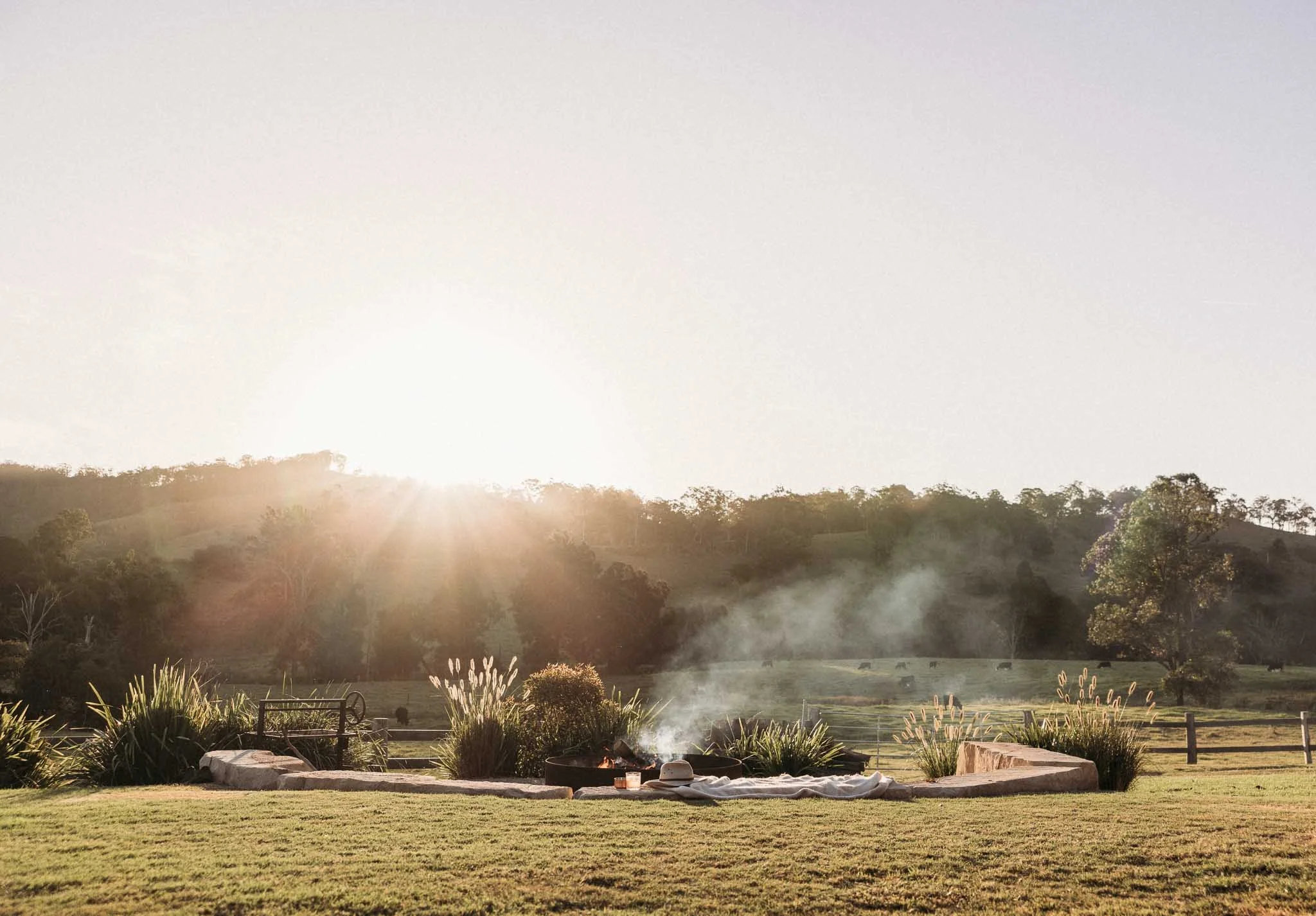 Sunset over a grassy lawn with trees and a fenced pasture, with smoke rising near a fire pit in the foreground.