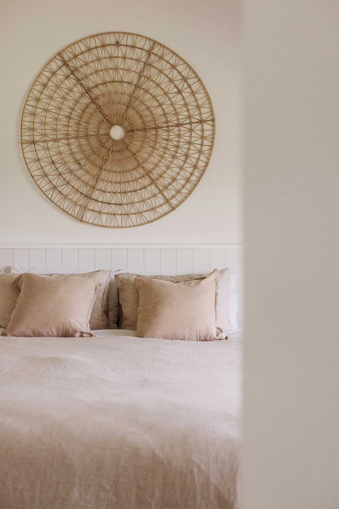 A bedroom wall decorated with a large circular woven wall hanging above a bed with beige pillows and a beige bedspread.