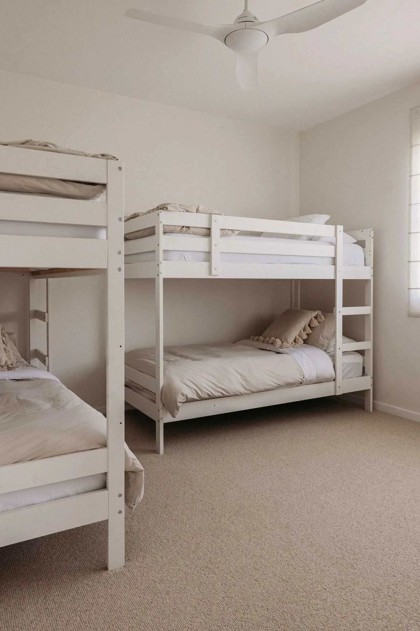 A bedroom with three white bunk beds and light-colored bedding and pillows, beige carpet, white walls, and a white ceiling fan.
