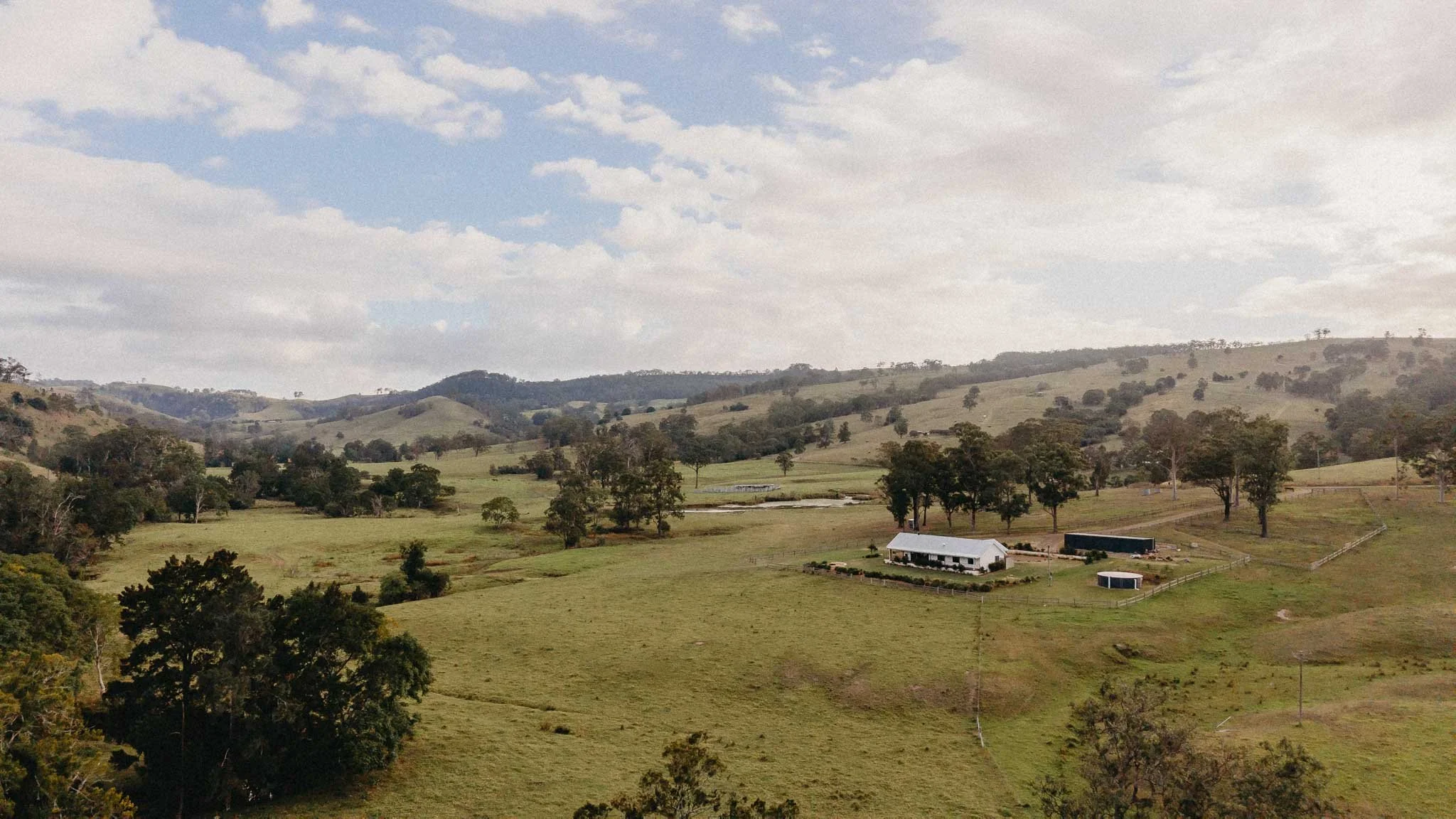 Scenic landscape of rolling green hills with scattered trees, a farm building with a white roof, and a cloudy sky overhead.