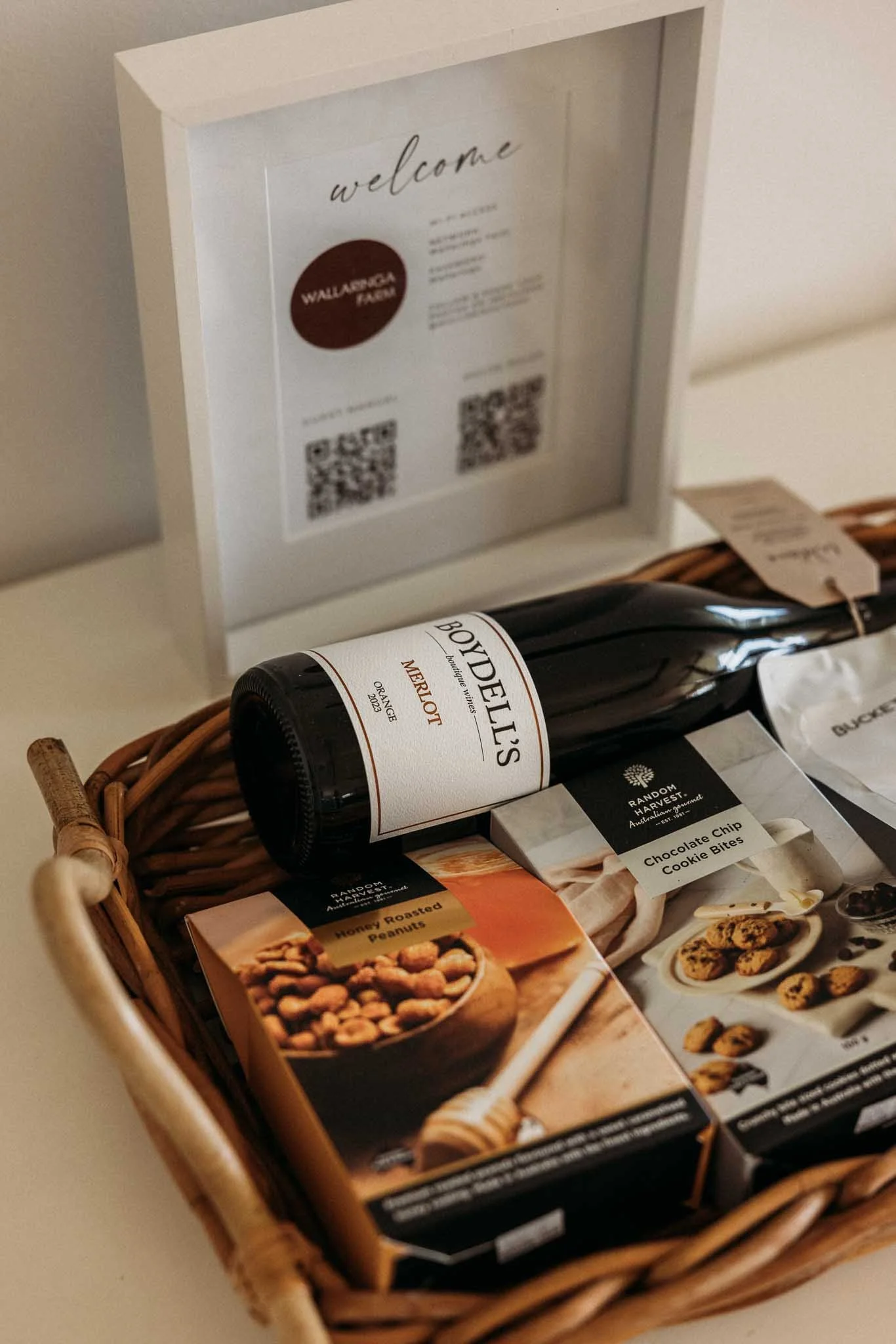 A wicker basket containing assorted snacks, including a bottle of Merlot wine, a box of honey roasted peanuts, and a box of chocolate chip cookie bites, with a white framed welcome sign in the background.