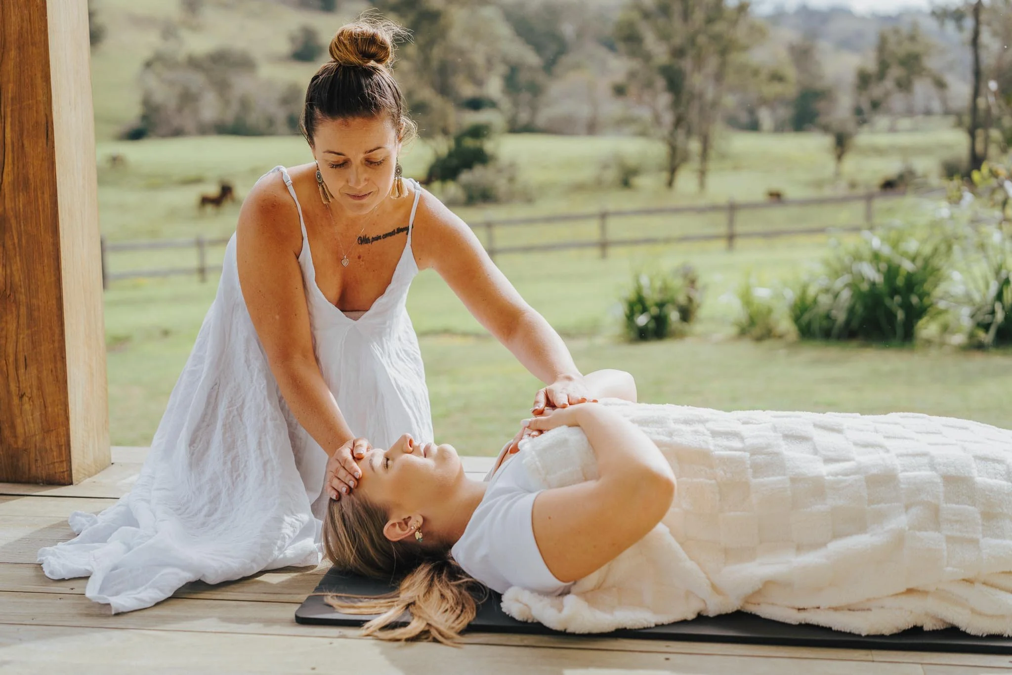 Woman lying on her back on a massage table outside, receiving a massage from another woman in a white dress, with a scenic countryside in the background.