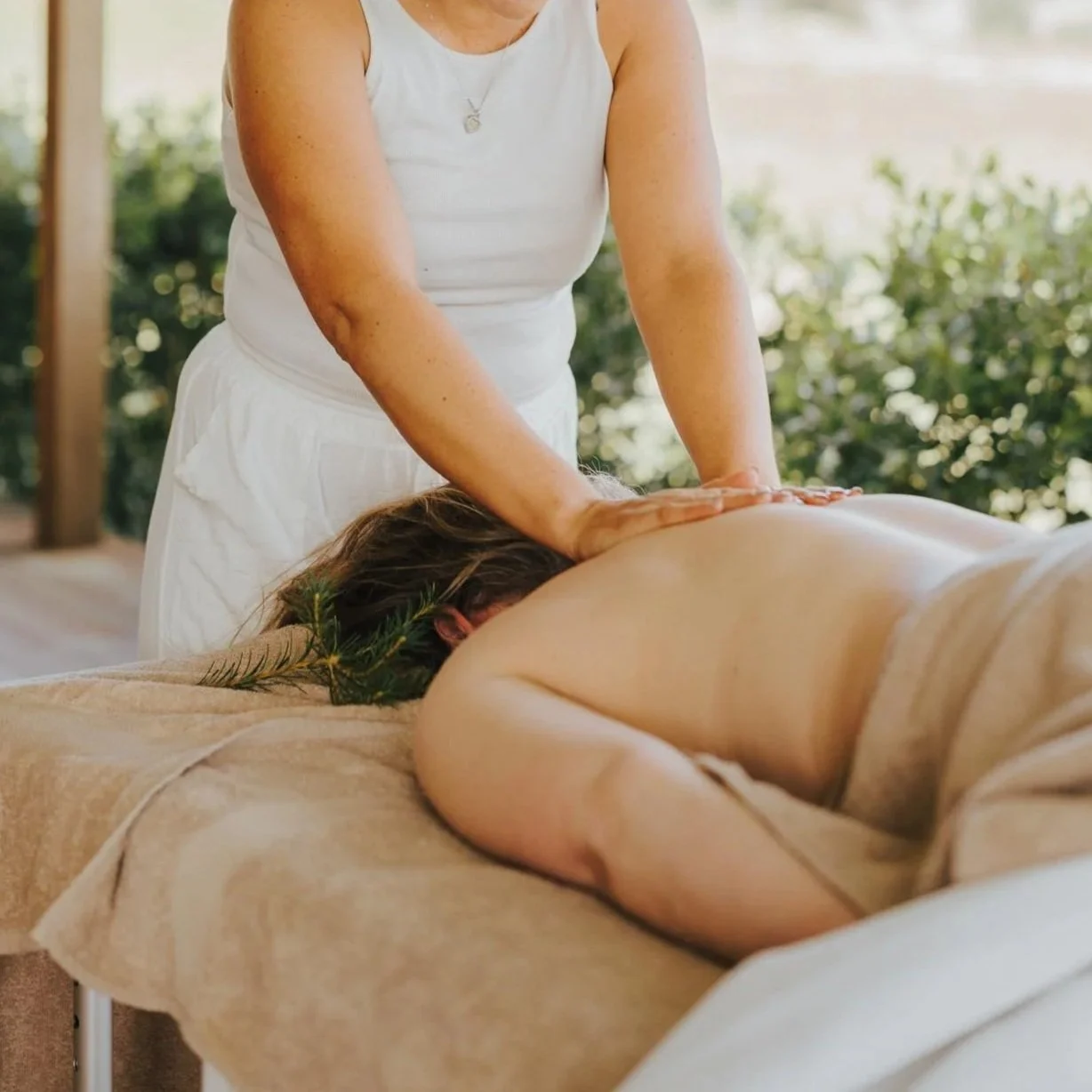 A person receiving a massage outdoors, lying face down on a massage table, with a massage therapist applying pressure to their back.