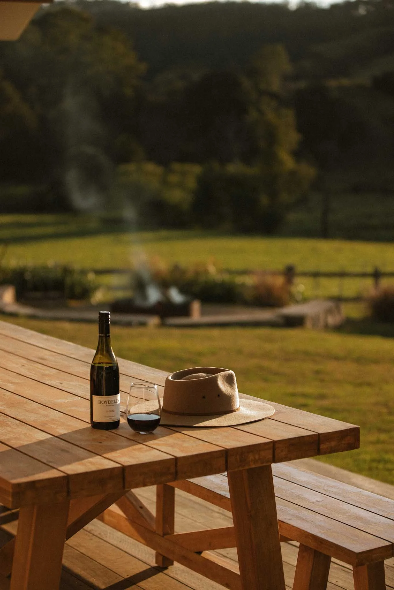 A wooden outdoor table set with a bottle of red wine, a glass of wine, a beige hat, and a cup, with a view of a grassy field and trees in the background at night.