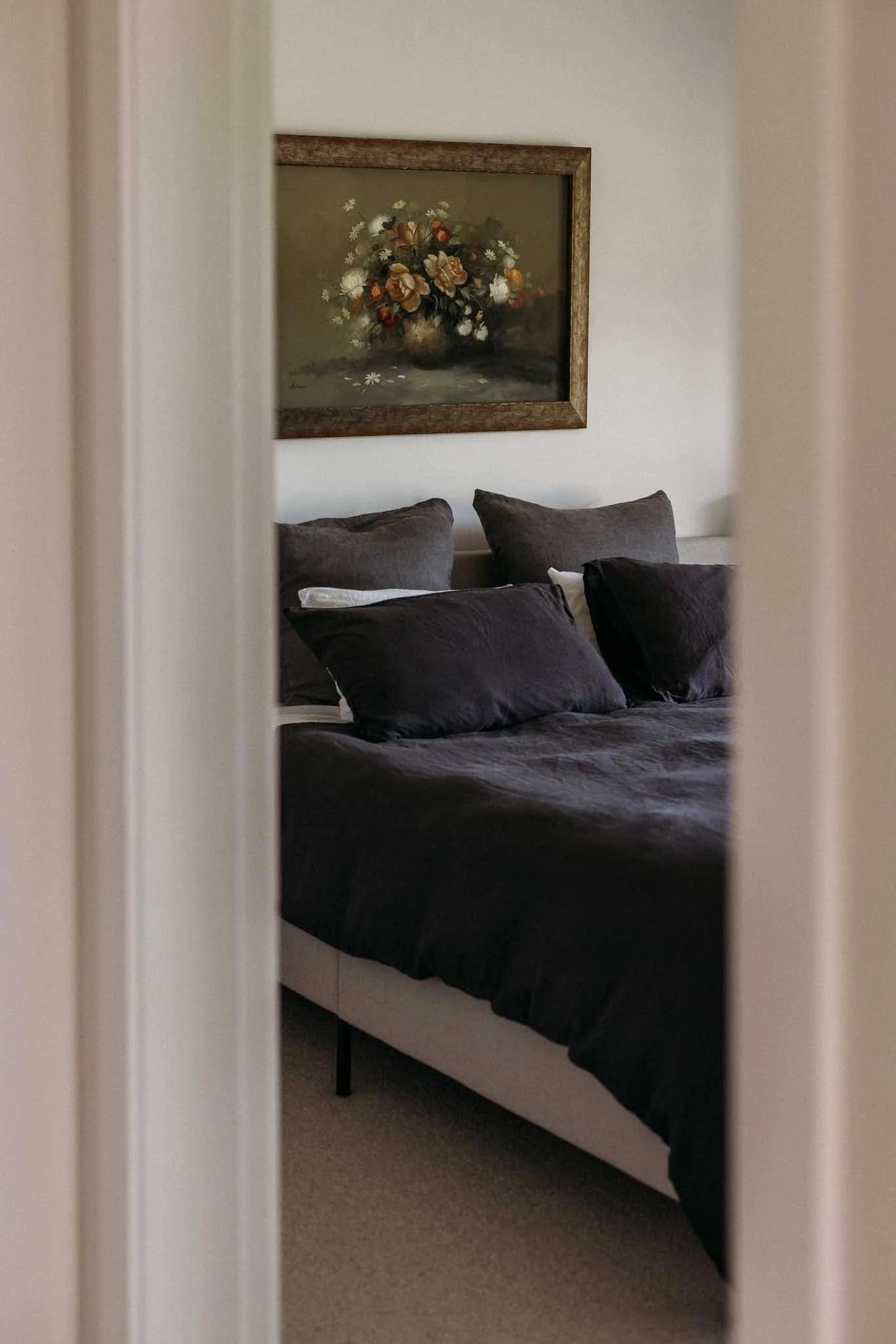 A bedroom with a bed, dark bedding, multiple pillows, a framed floral painting, and a carpeted floor, viewed through a partially open door.