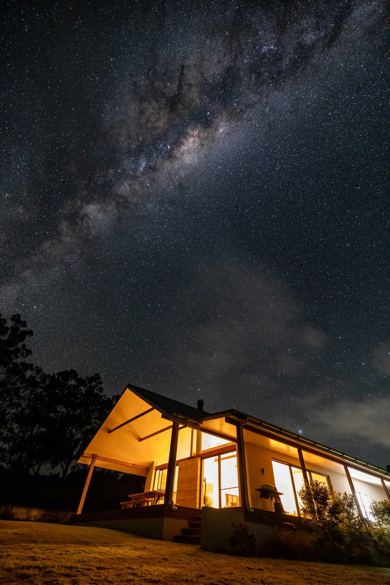 Modern house with warm interior lighting under a starry night sky, including the Milky Way galaxy, with trees and a lawn in the foreground.