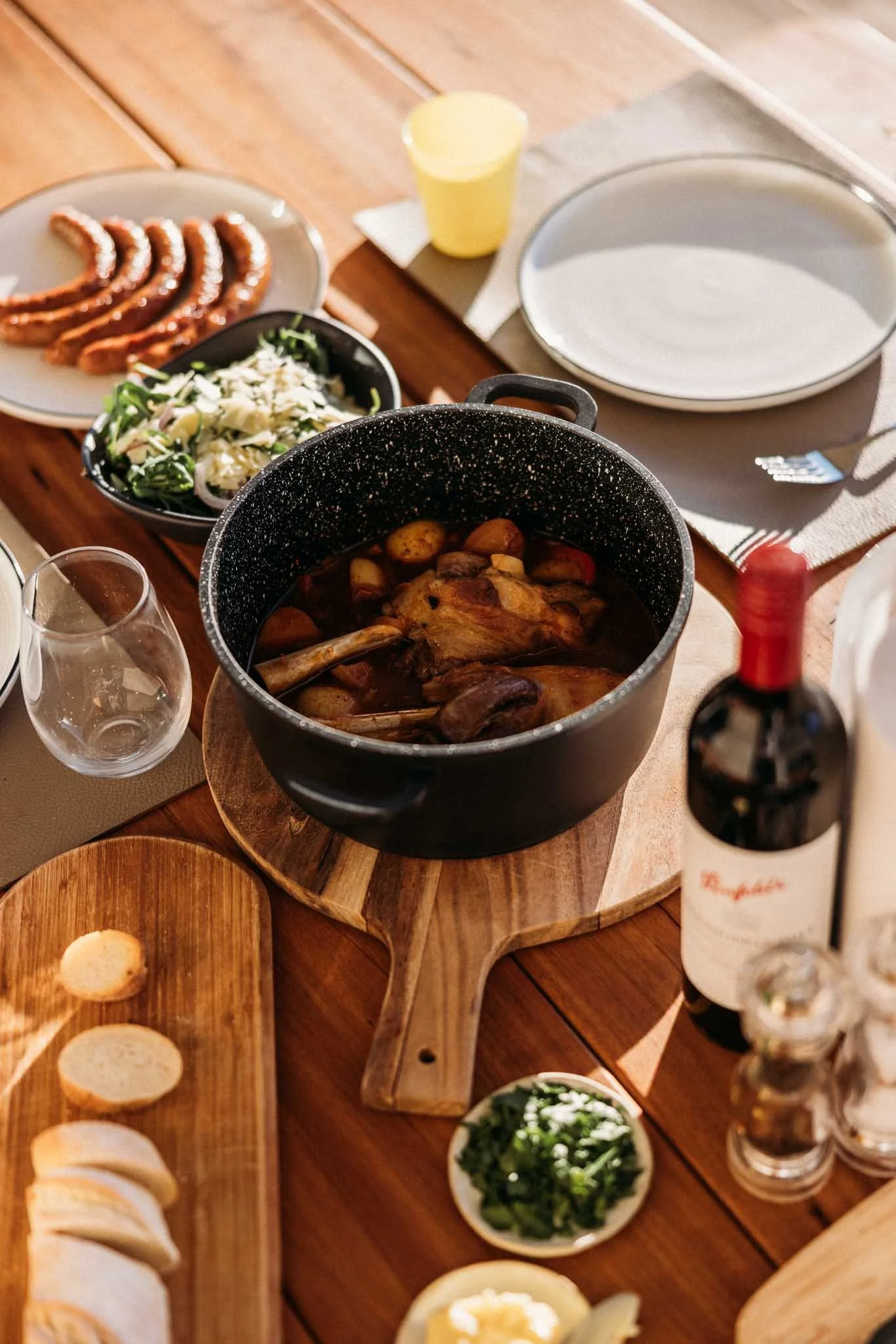 A rustic wooden table set with various dishes including a pot of cooked meat, sliced bread on a wooden board, a salad, curly fries, a glass of water, and a bottle of red wine.