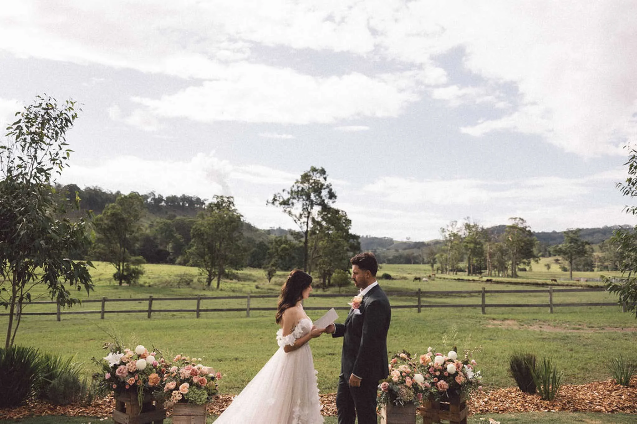 A bride and groom exchanging vows outdoors in a rural setting with trees, a grassy field, and hills in the background. The bride wears a white wedding dress with a long train, and the groom is in a dark suit. They stand under a floral arch with pink and white roses, with a wooden fence at the back.
