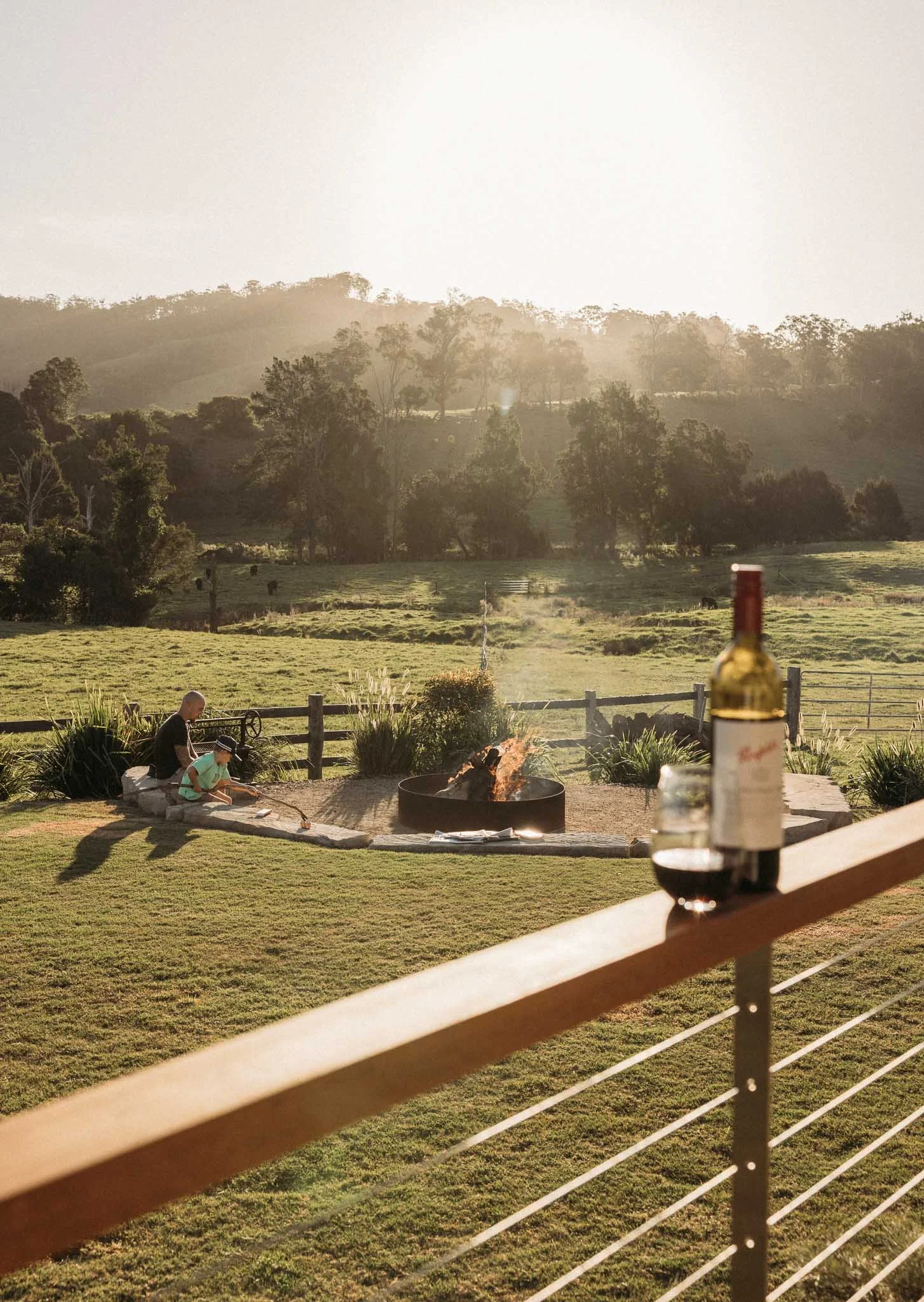 A backyard scene with two children sitting by a fire, a fence in the foreground, a bottle of wine, and a green field with trees and hills in the background at sunset.