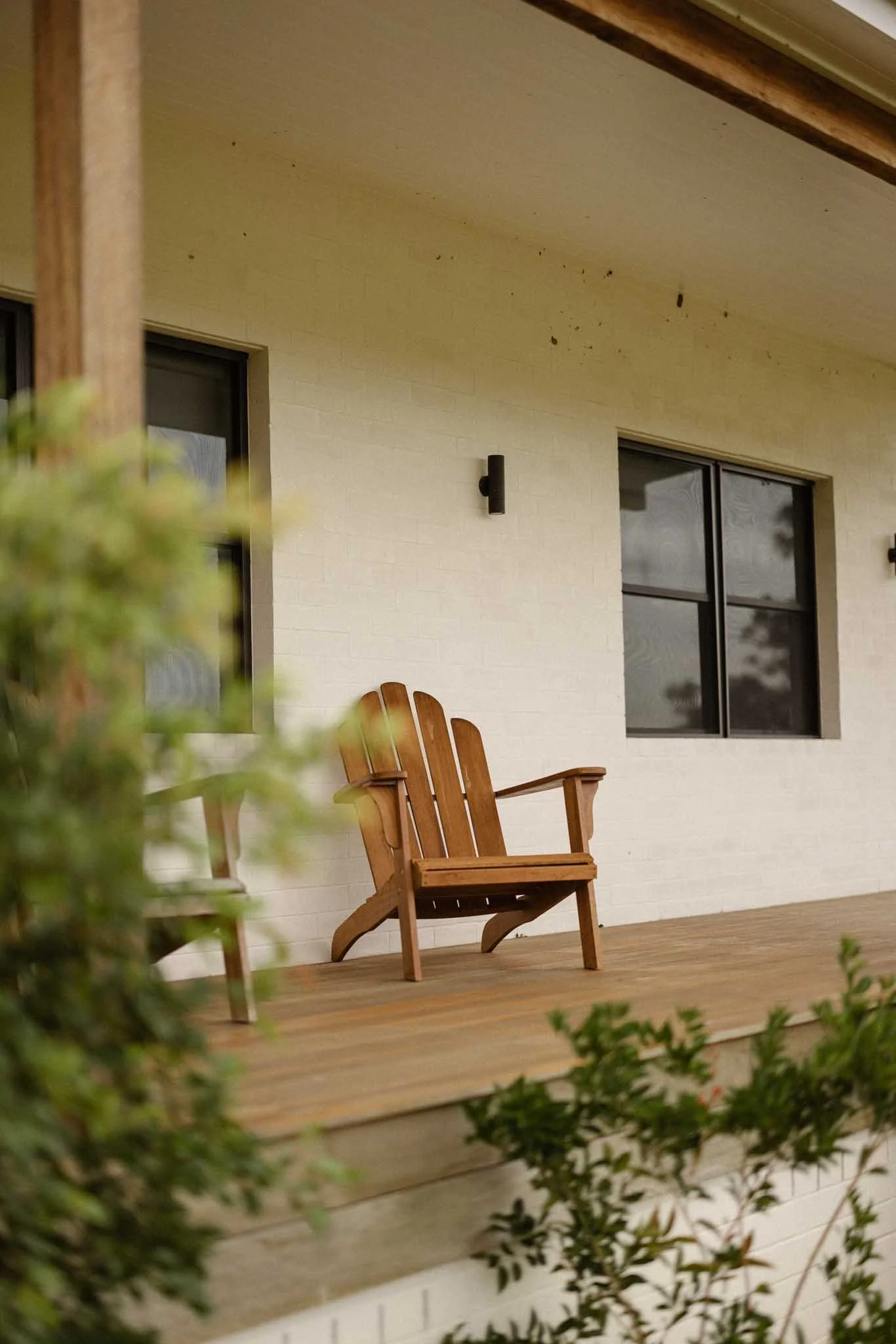 A wooden chair on a porch with a white brick wall, two black windowpanes, and a light fixture. Green plants frame the foreground.
