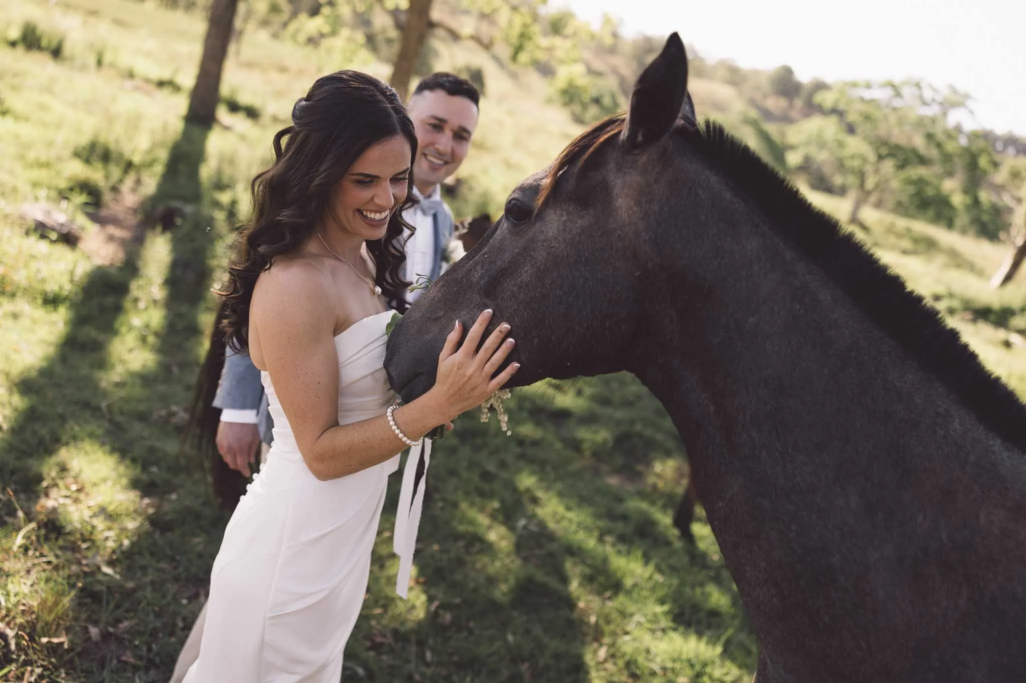 A woman in a white dress smiling and petting a dark horse on its face outdoors with trees and sunlight in the background, accompanied by a man in a suit behind her.