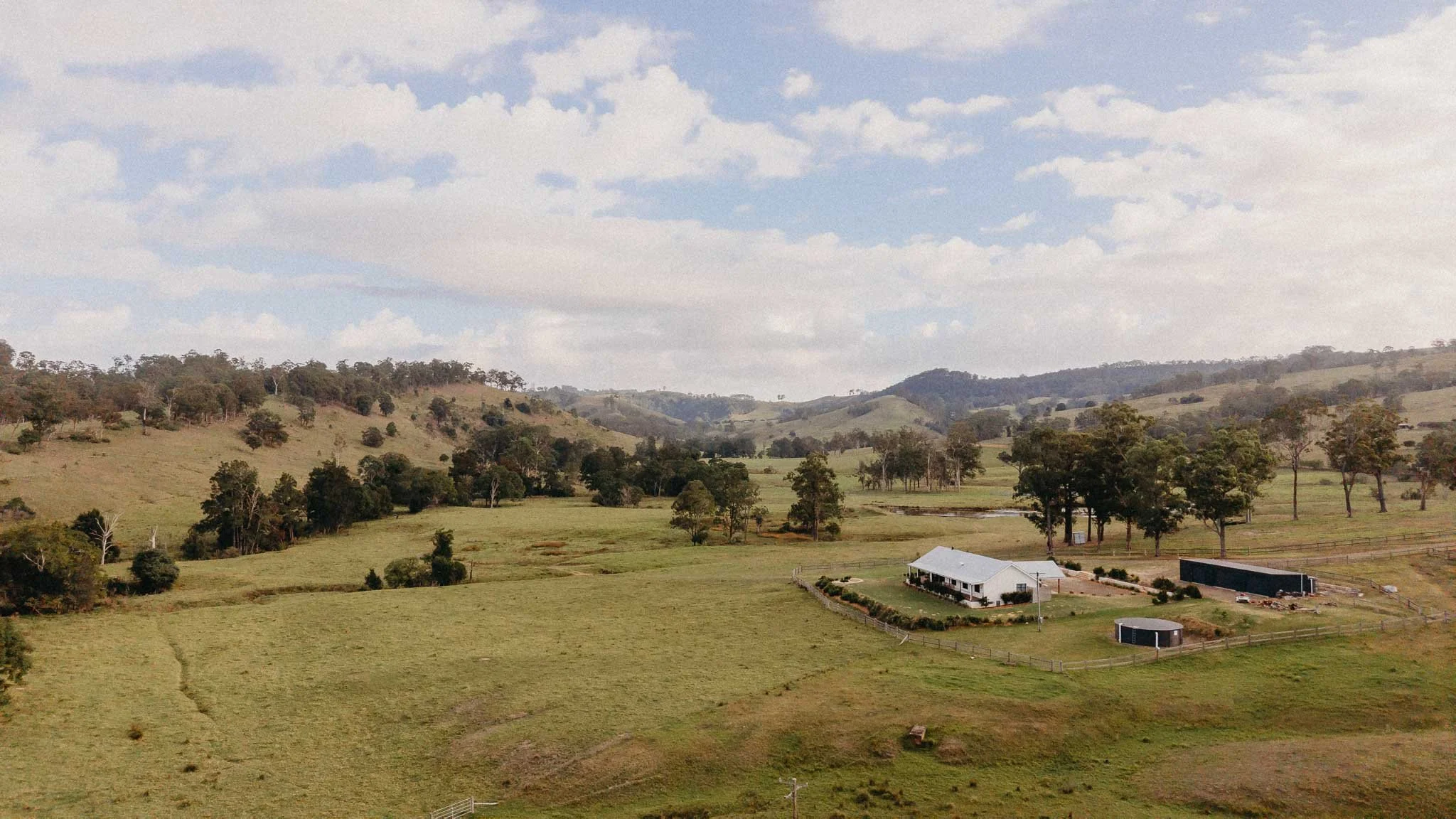 A rural landscape with green rolling hills, scattered trees, and a cloudy sky. There is a white house with a porch, a shed, and a fenced yard in the foreground.