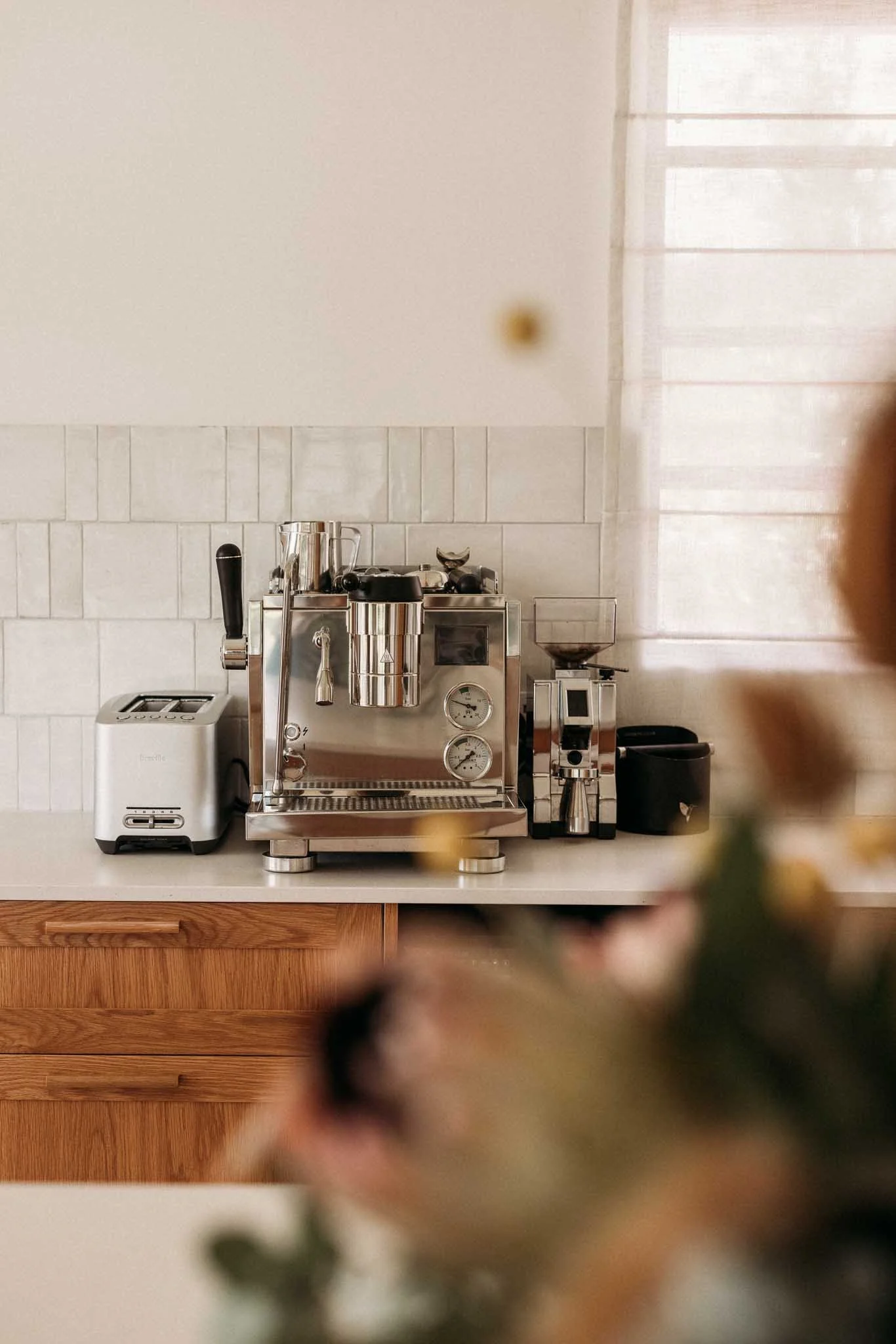 A kitchen countertop with a white toaster, a high-efficiency espresso machine, and a coffee grinder, with a tiled backsplash and a window with sheer curtains.