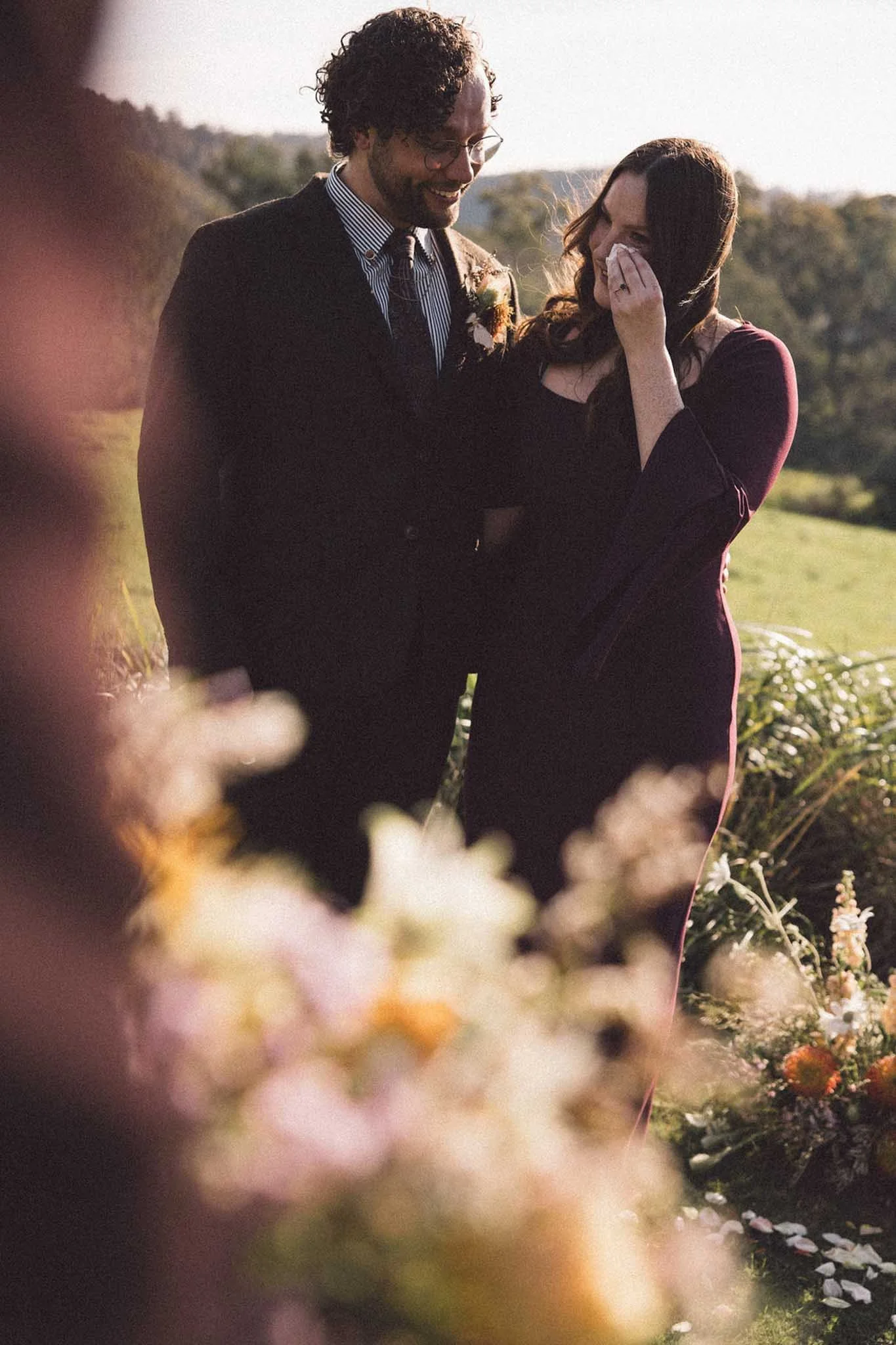 A man and woman share a joyful moment outdoors, surrounded by flowers and greenery, with the woman wiping away tears of happiness.