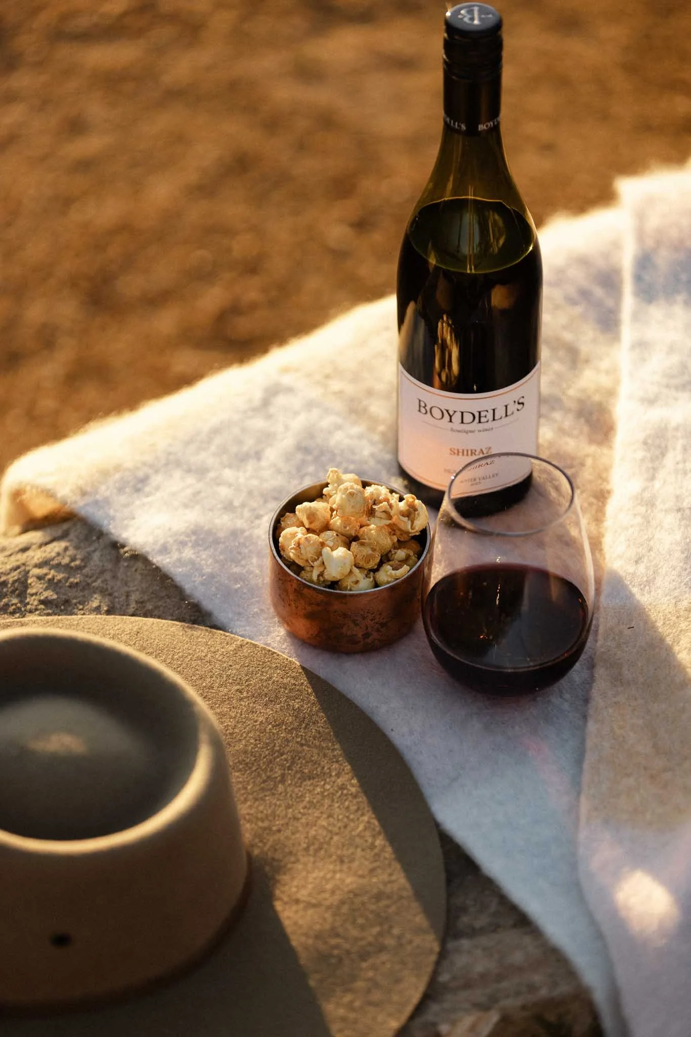 A bottle of Boydell's Shiraz wine, a glass of red wine, a small bowl of popcorn, and a beige cup on a table with a white cloth and a brown surface underneath.