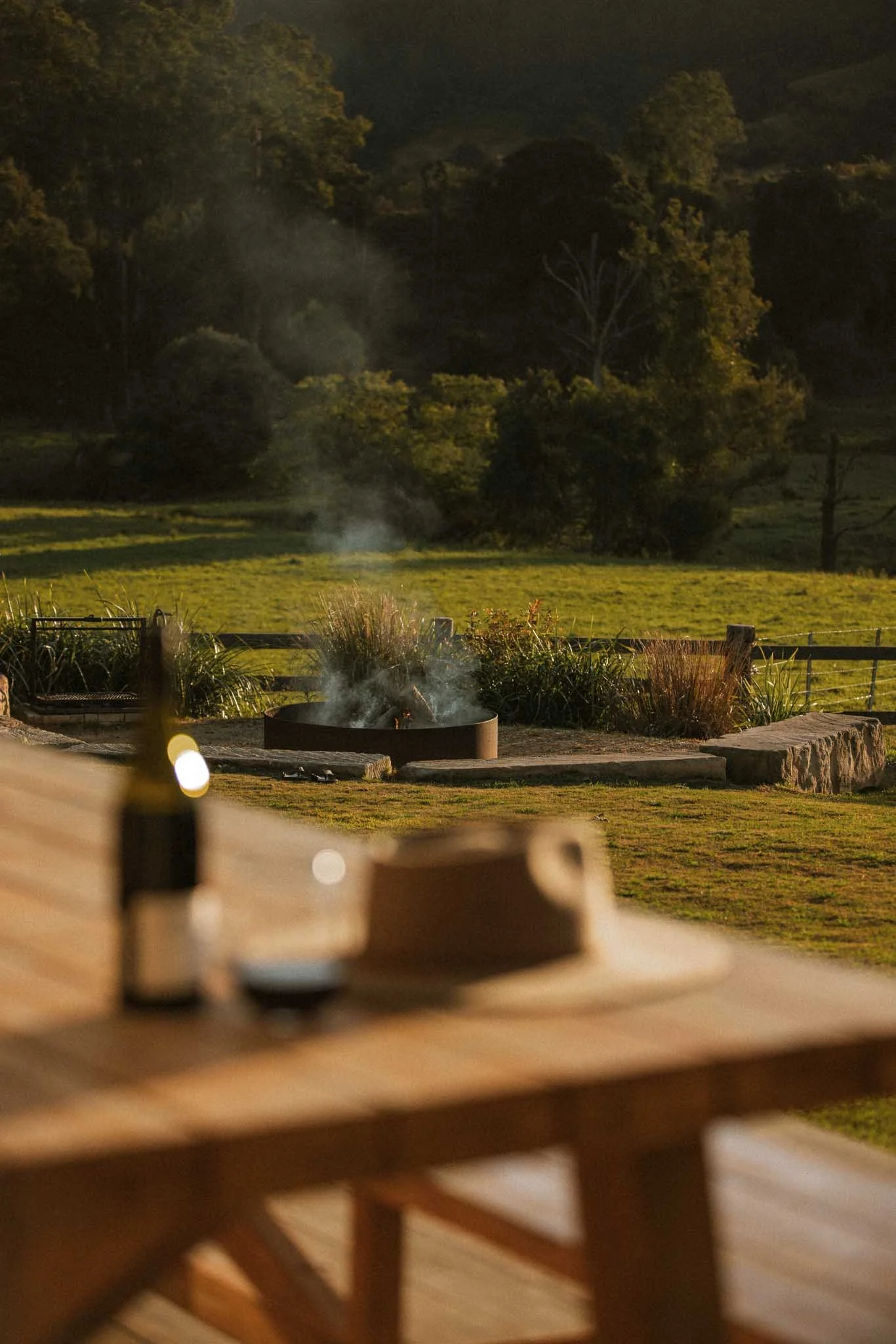 Nighttime outdoor scene with a fire pit emitting smoke, surrounded by a grassy area, trees, and hills in the background. The photo is taken from a wooden deck with a railing in the foreground.