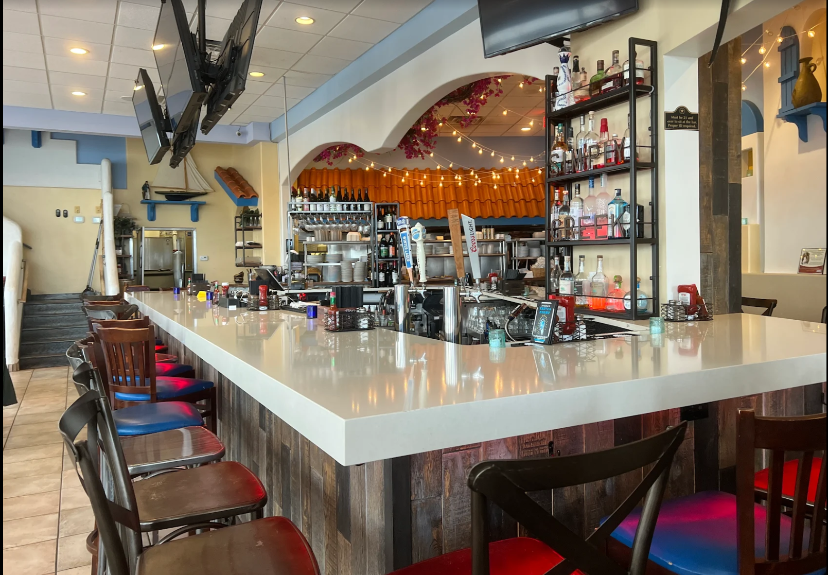 Interior of a bar, featuring a white counter with barstools, a back shelf with liquor bottles, and bar equipment. There are multiple TV screens hanging from the ceiling, and decorative string lights are visible in the background.
