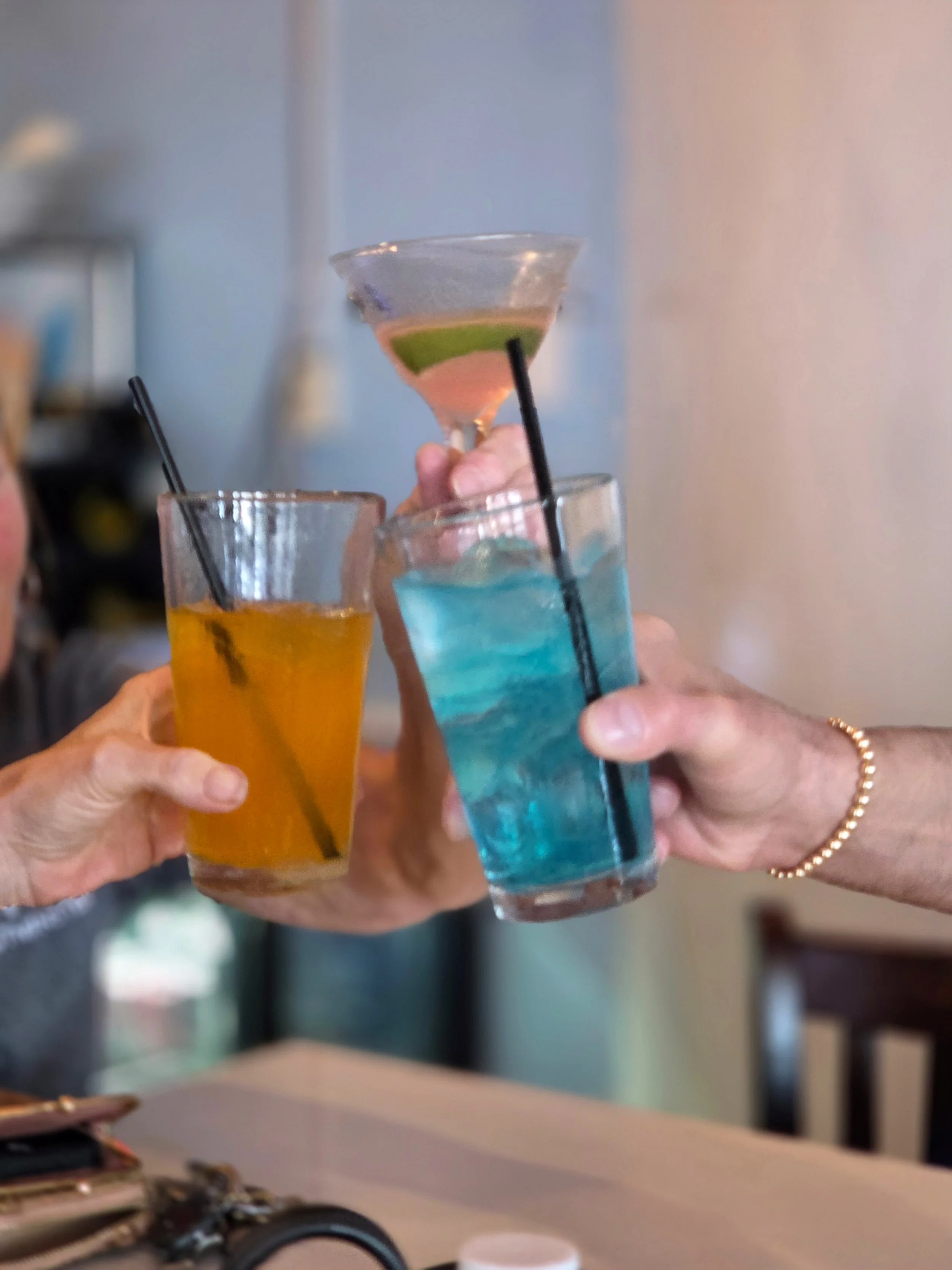 Multiple hands holding colorful cocktails with straws, raising glasses together in a toast.