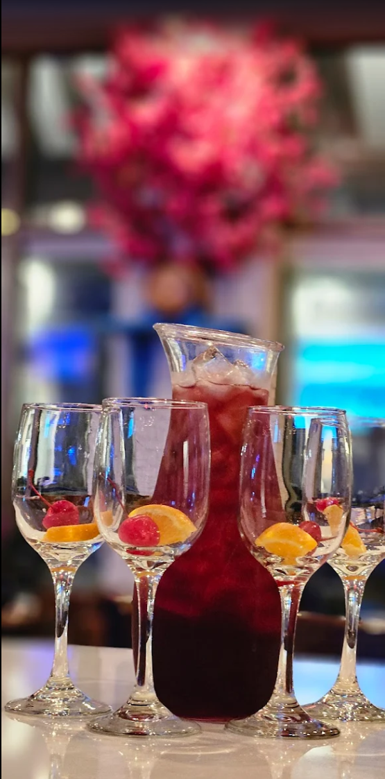 A pitcher of red sangria with ice, garnished with cherries and orange slices, surrounded by four empty wine glasses with cherries and orange slices inside, set on a table at a social event, with a blurred floral arrangement in the background.