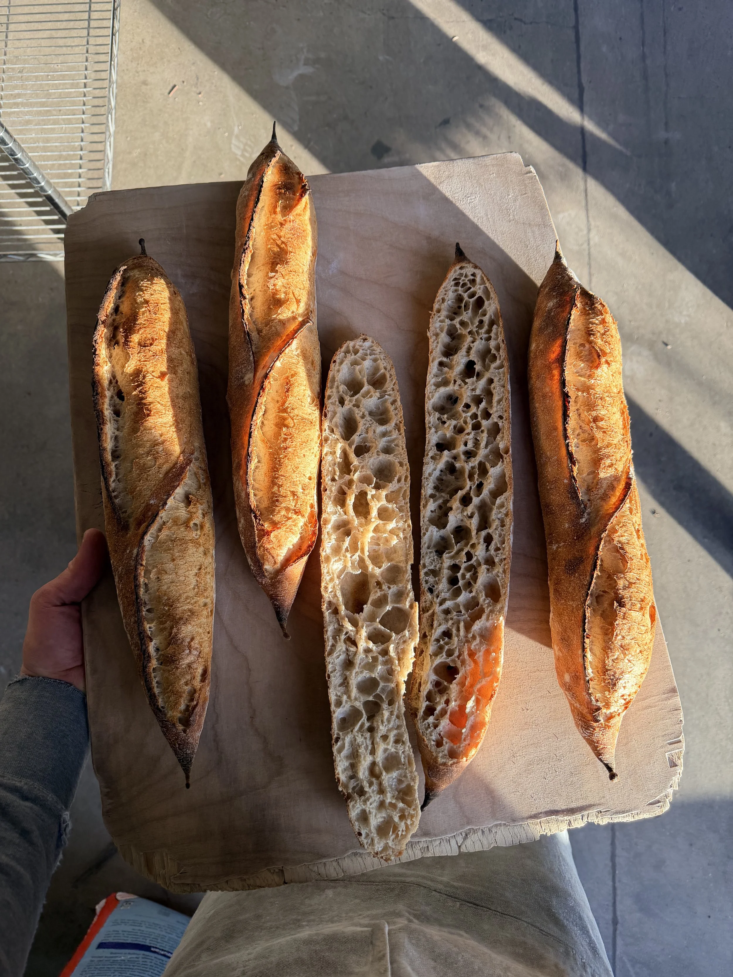 Five freshly baked baguettes with a golden crust and open crumb, resting on a wooden board.