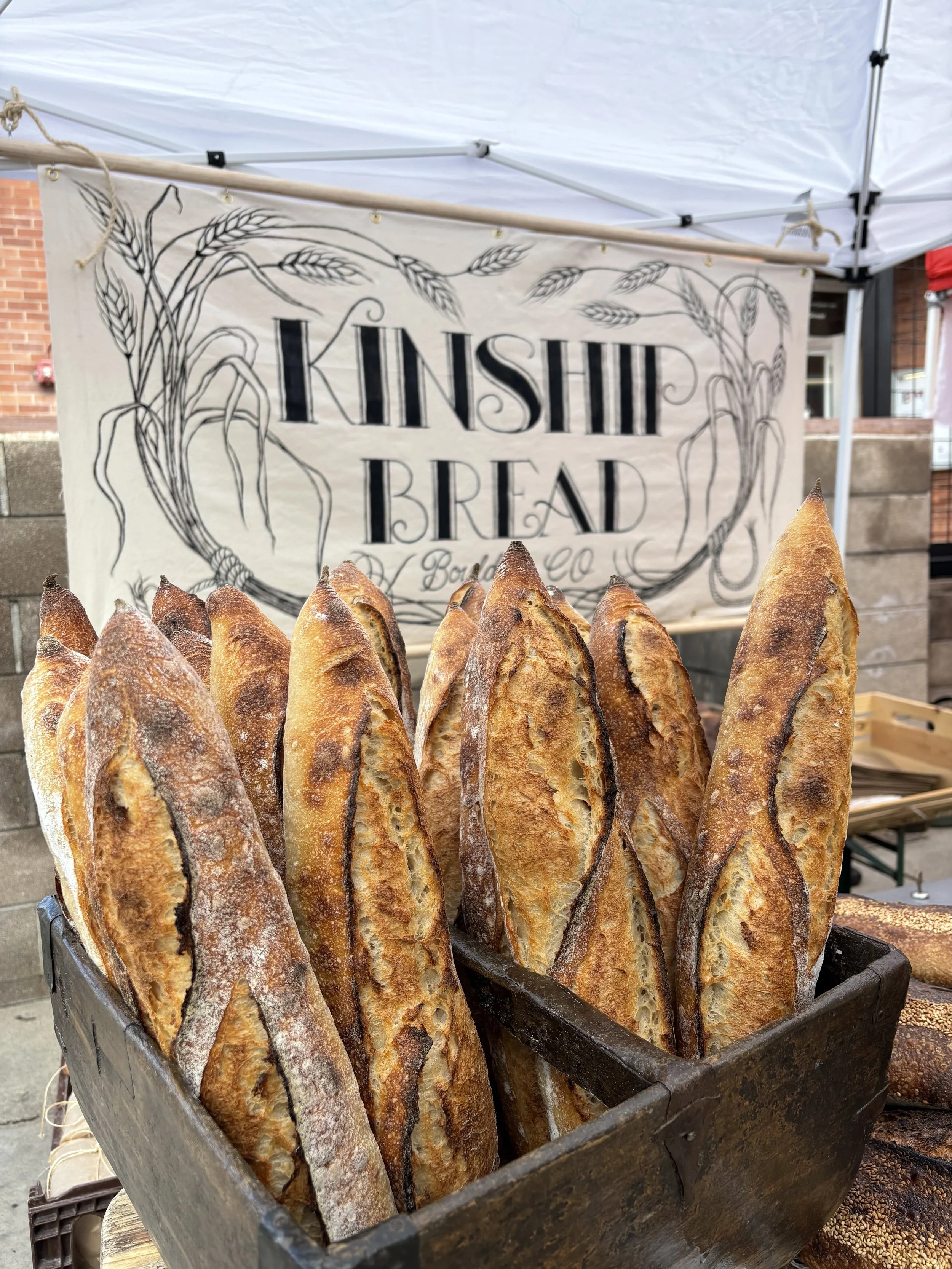 A wooden basket filled with freshly baked baguette loaves at an outdoor market stall with a sign that reads 'Kinsch Bread' in the background.