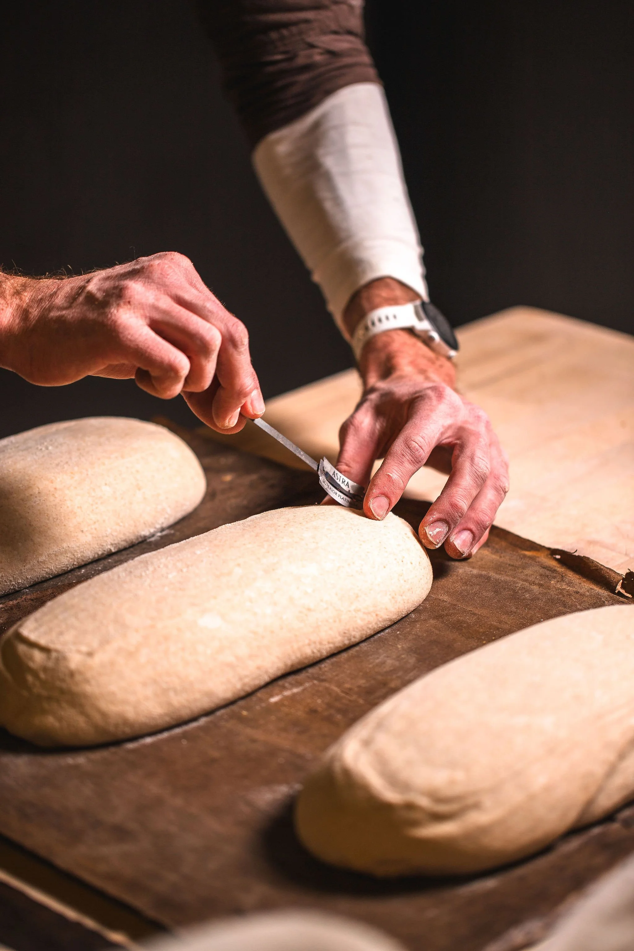 A person with a bandaged arm is carefully cutting into loaves of freshly baked bread on a wooden table.