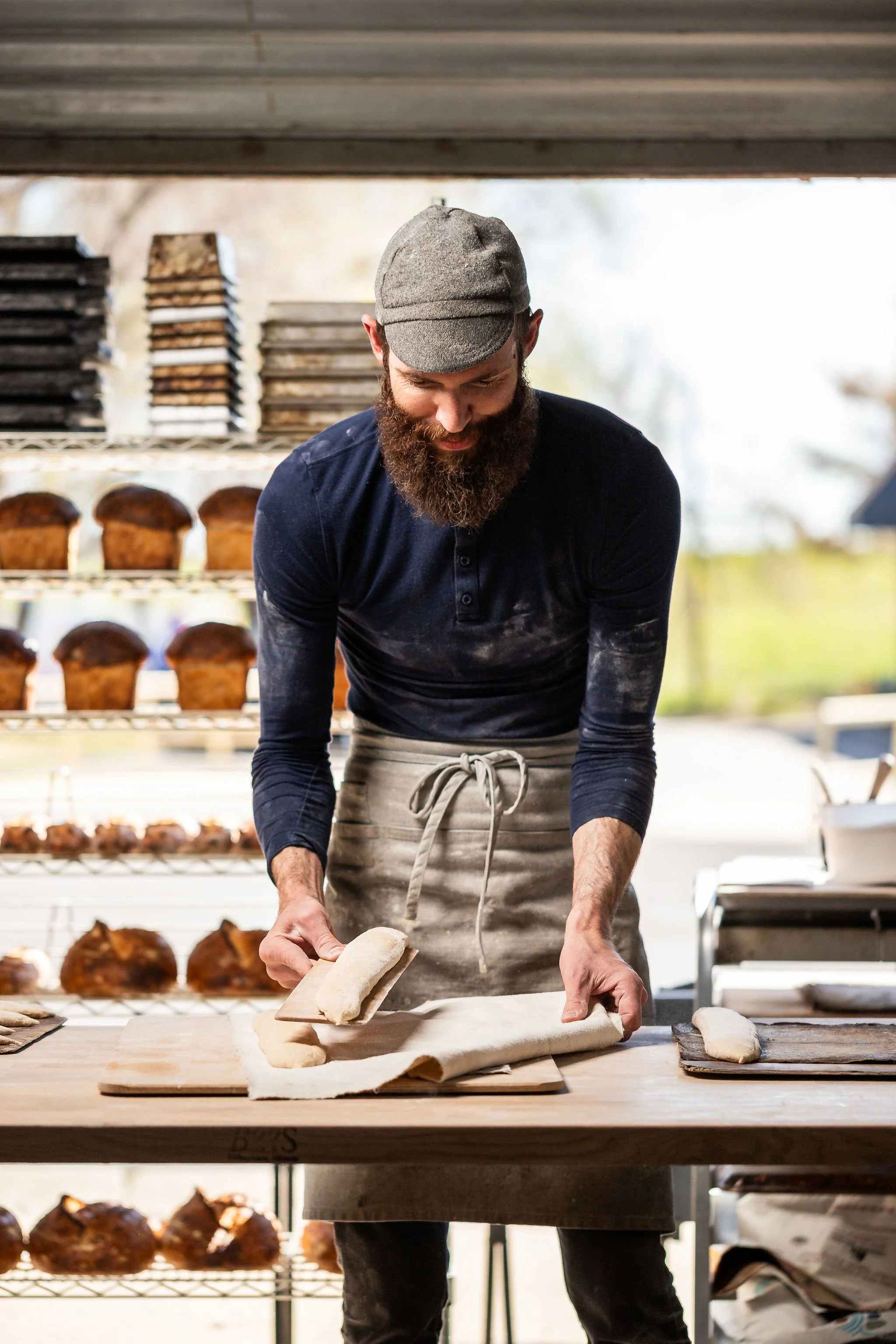 A man with a beard and gray cap preparing dough on a wooden surface in a bakery, with shelves of baked goods in the background.