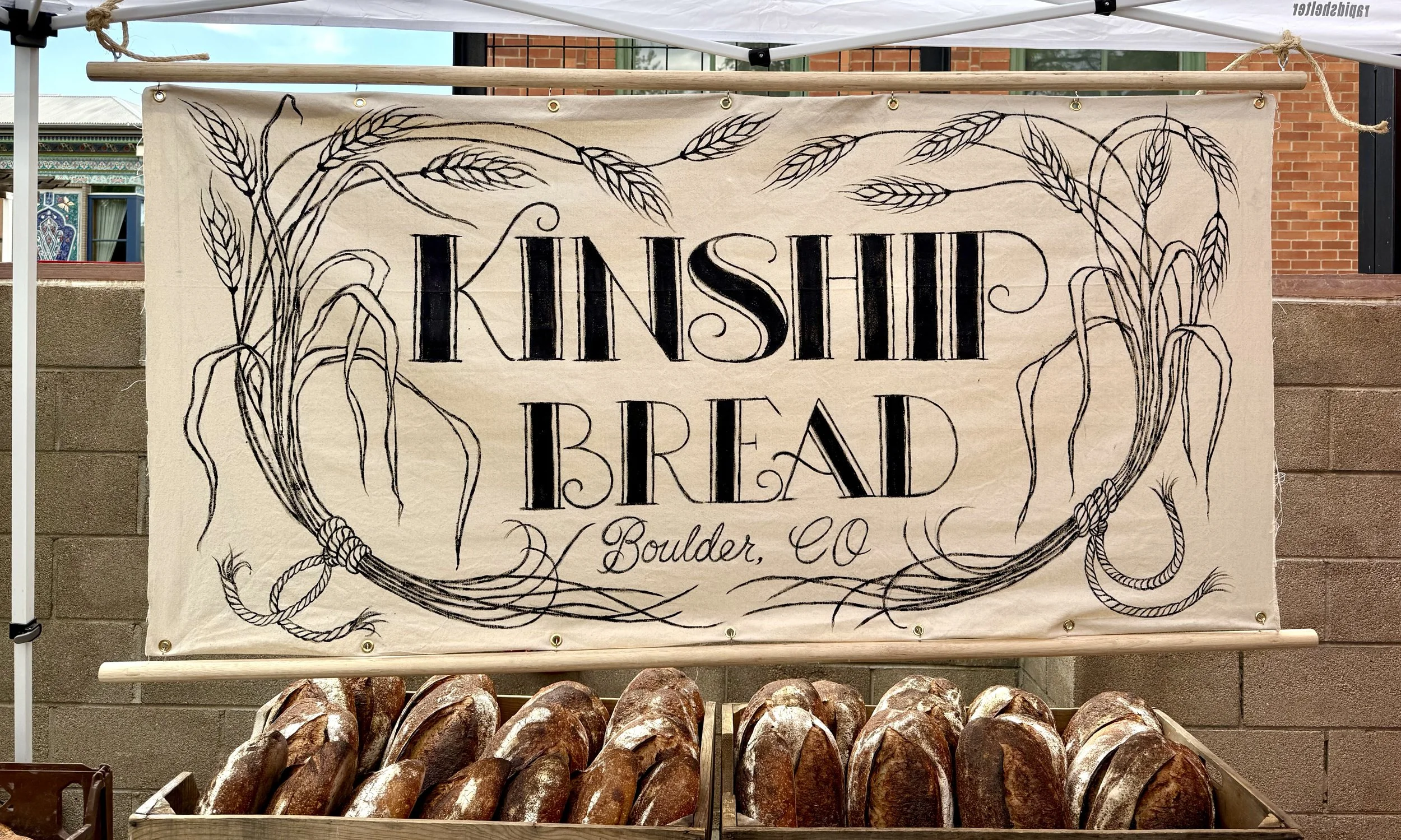 A fabric banner with the words 'Kinship Bread' and 'Boulder, CO' written on it, decorated with illustrations of wheat and tied with rope at the corners. Bread loaves are stacked below the banner.