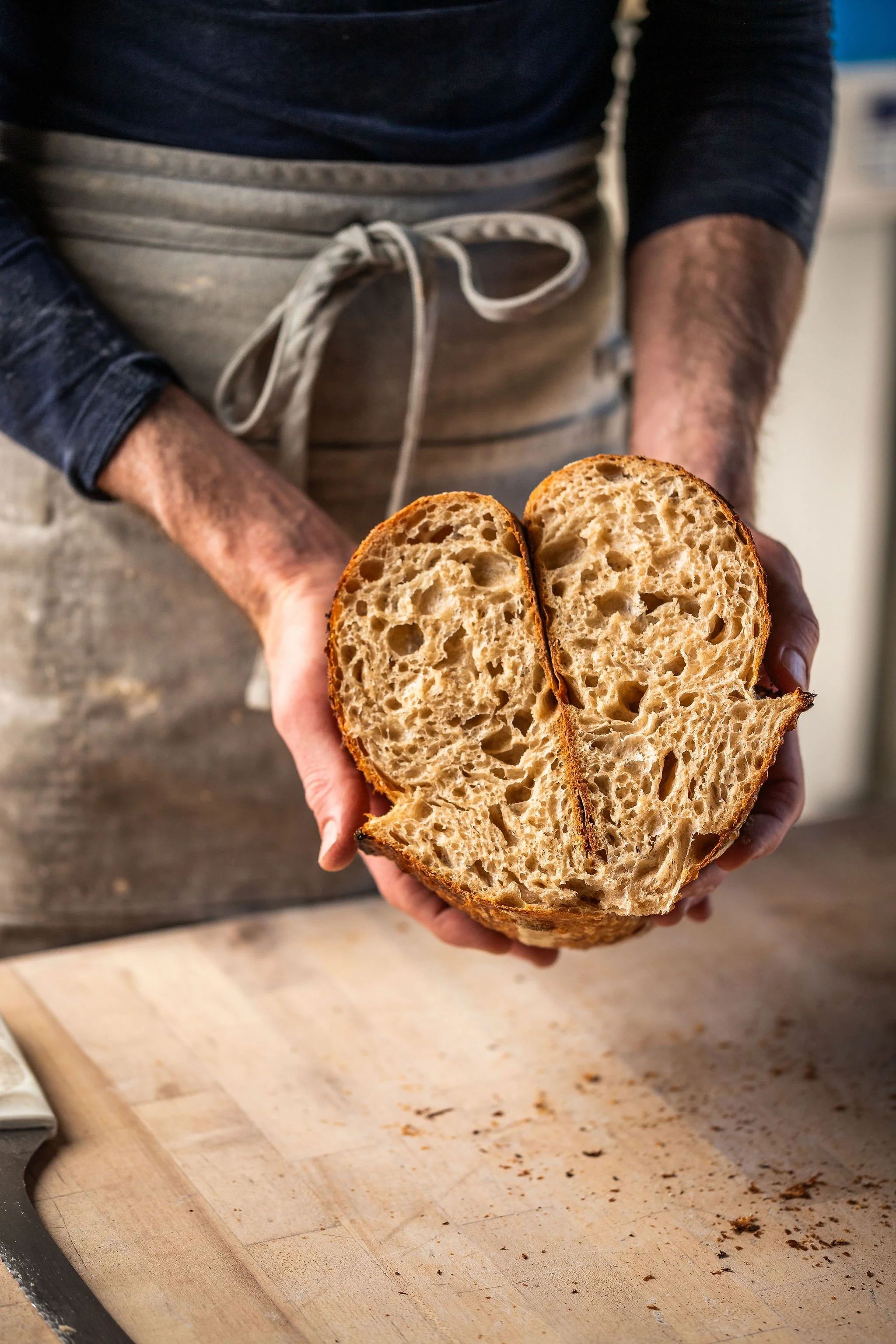 A person wearing a gray apron and a dark long-sleeve shirt holds a halved loaf of freshly baked bread. The loaf has a perforated, airy interior and a crusty outer layer. The person is standing in a kitchen with a wooden cutting board that has crumbs on it.