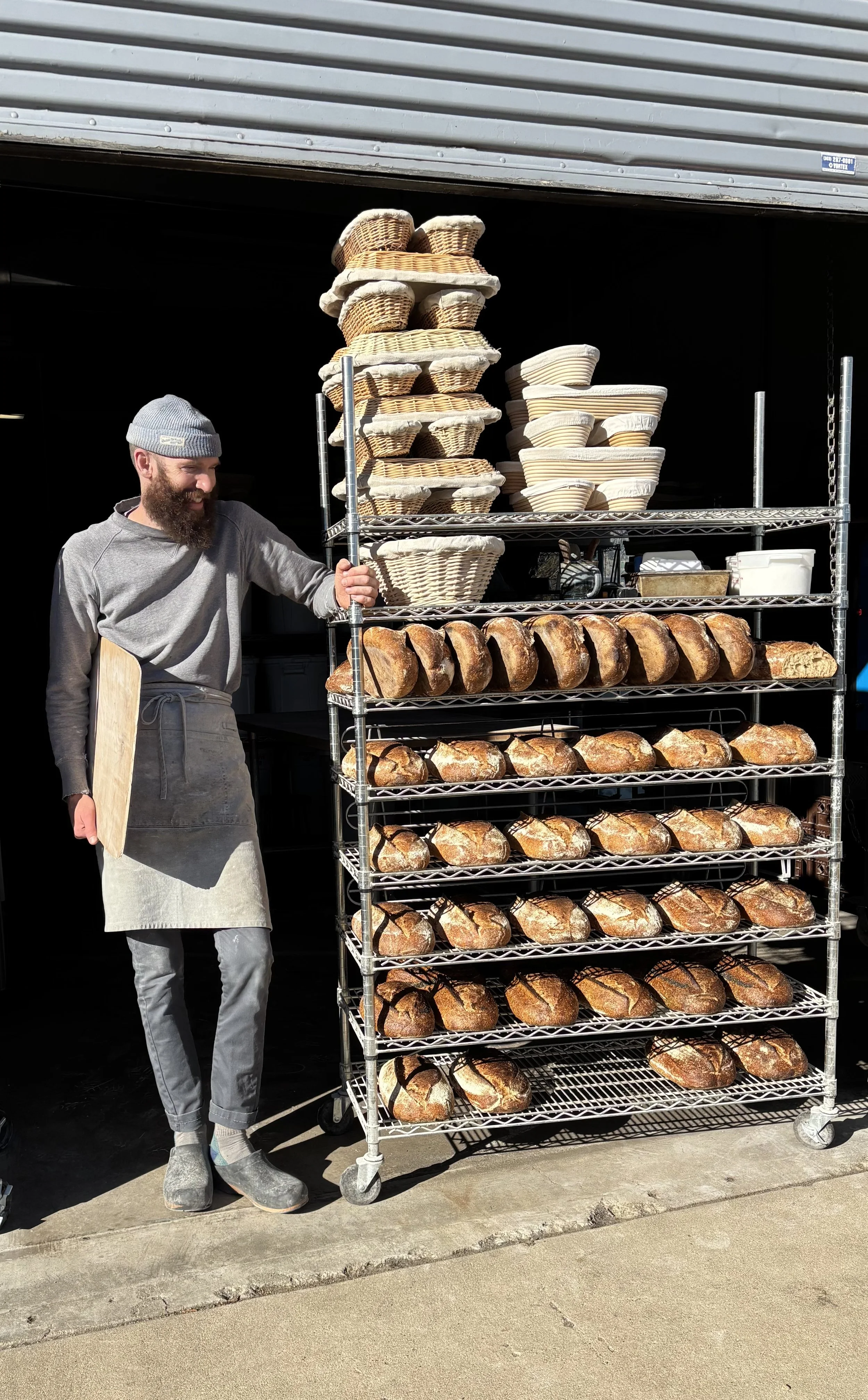 A man with a beard, gray beanie, gray shirt, apron, and boots standing next to a large metal bakery rack filled with freshly baked bread loaves.