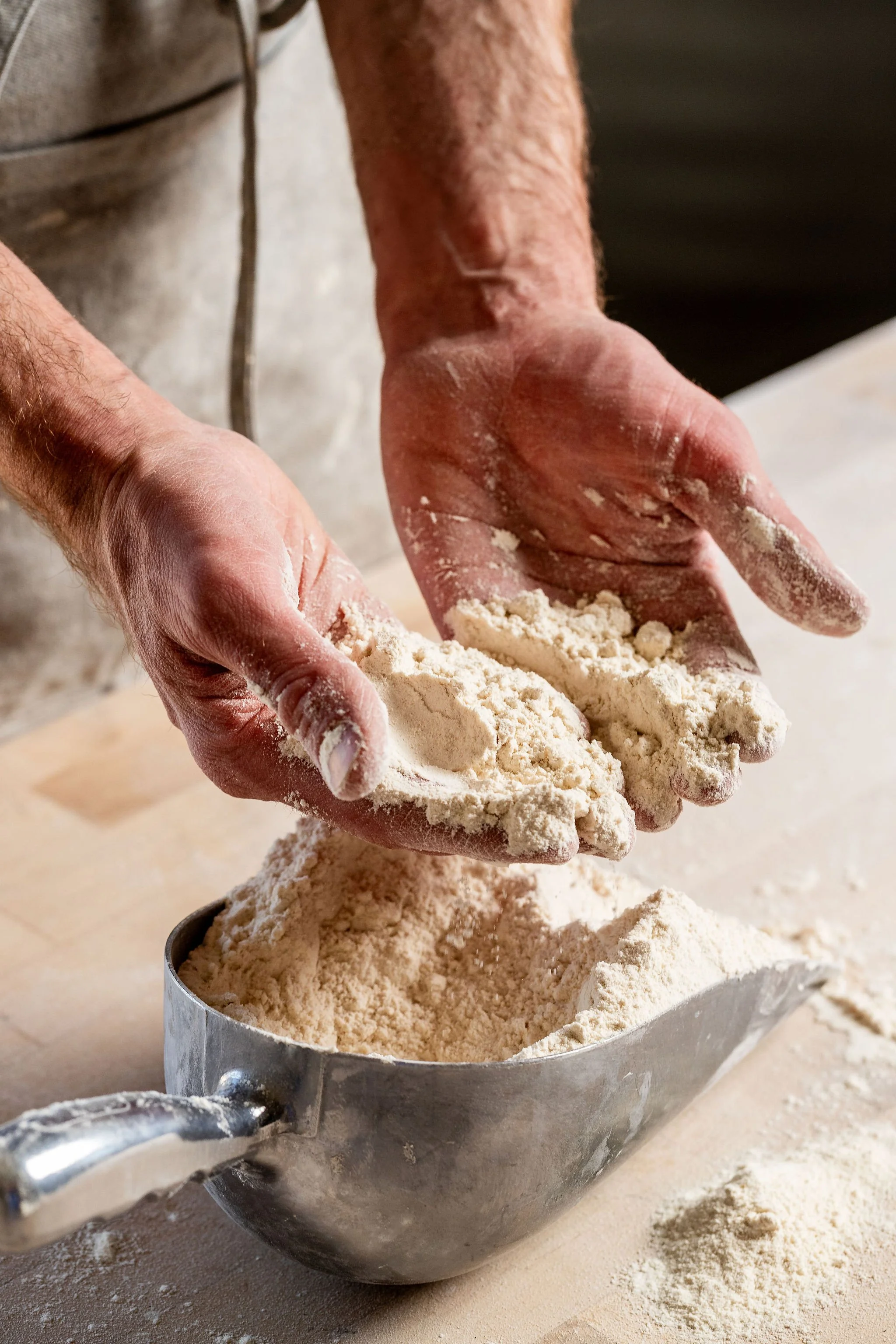 Close-up of hands sifting flour into a metal mixing bowl on a countertop, with some flour scattered around.
