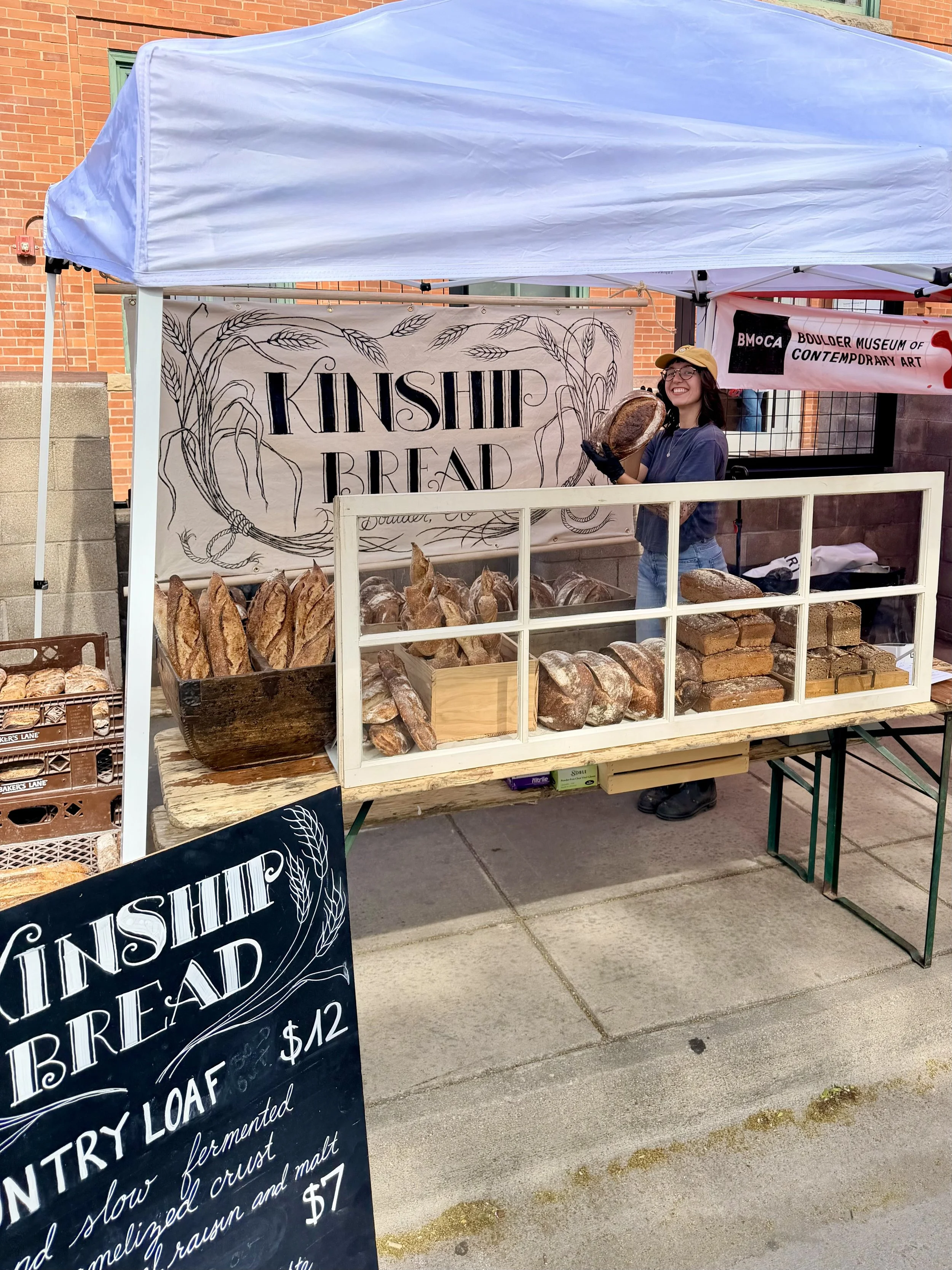 A woman standing behind a bread stall at an outdoor market, smiling and holding a loaf of bread. The stall has a sign reading "KINSHIP BREAD" with various loaves of bread displayed in front. There are signs for other items and prices nearby.