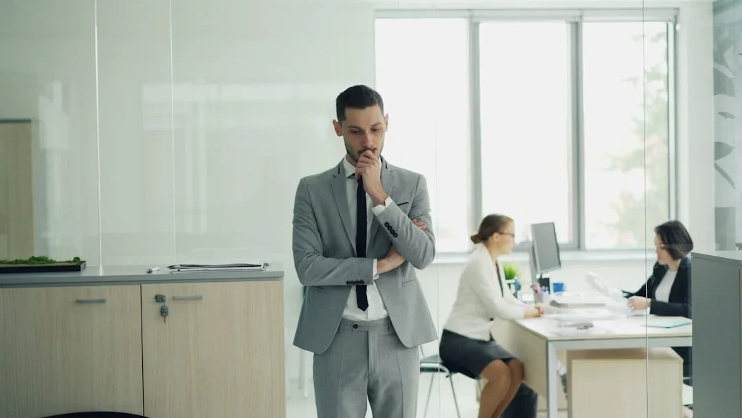 Un homme en costume gris, pensif, dans un bureau moderne avec deux femmes travaillant à leur bureau en arrière-plan.