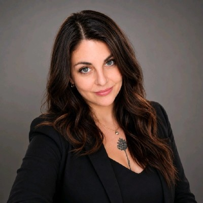 Une femme avec des cheveux bruns et longs, portant un blazer noir, souriant légèrement, avec un fond gris.